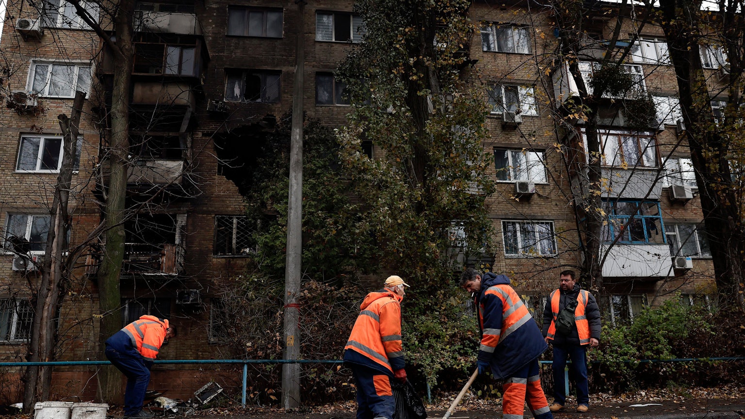 Arbeiter in orangen Westen räumen mit Schauefeln Schutteile eines Wohnhauses weg. Das Haus ist im Hintergrund zu sehen, in der Wand klafft ein großes, dunkles Loch nach einem Raketeneinschlag.