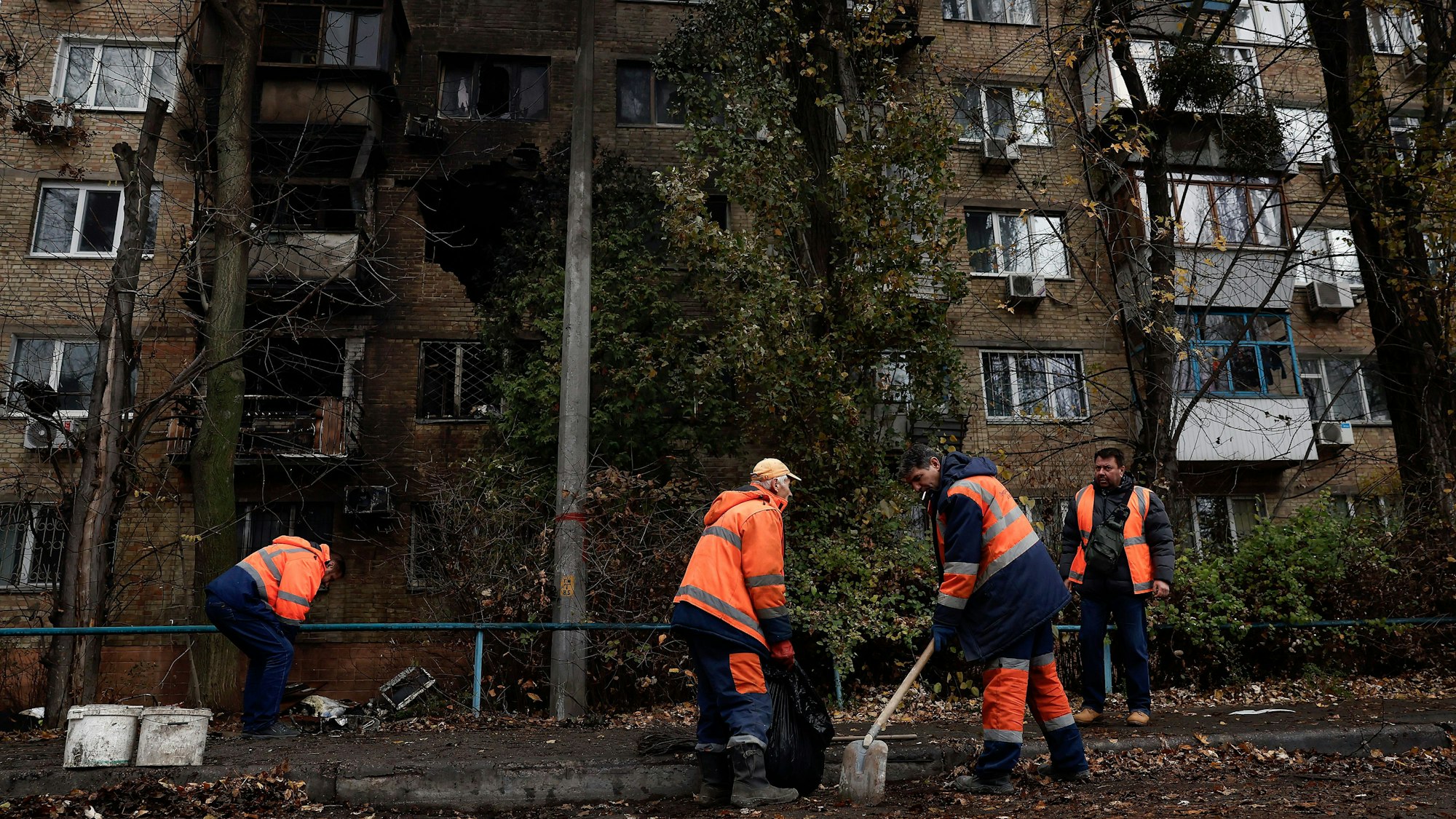 Arbeiter in orangen Westen räumen mit Schauefeln Schutteile eines Wohnhauses weg. Das Haus ist im Hintergrund zu sehen, in der Wand klafft ein großes, dunkles Loch nach einem Raketeneinschlag.