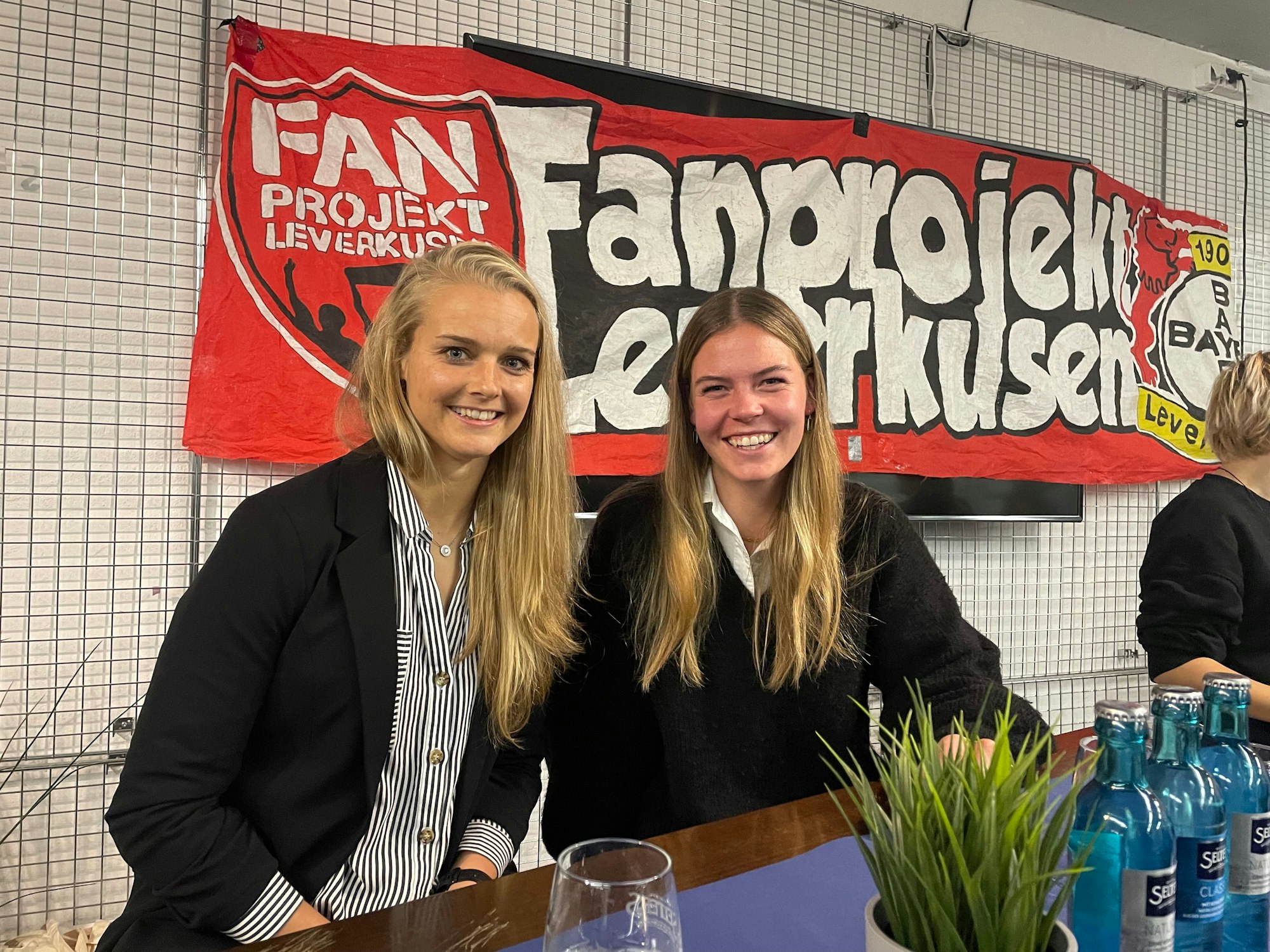 Die Spielerinnen Anna Klink und Juliane Wirtz von Bayer 04 Leverkusen. Im Hintergrund hängt ein Banner des Fanprojekts Leverkusen an der Wand.