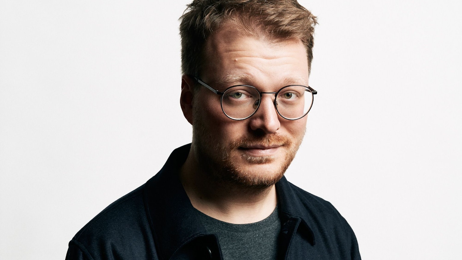 Stand-Up-Comedian Maxi Gstettenbauer steht vor einer weißen Wand. Er trägt ein blaues Hemd über einem grauen T-Shirt.