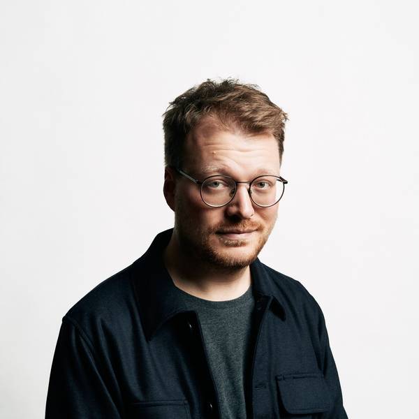 Stand-Up-Comedian Maxi Gstettenbauer steht vor einer weißen Wand. Er trägt ein blaues Hemd über einem grauen T-Shirt.