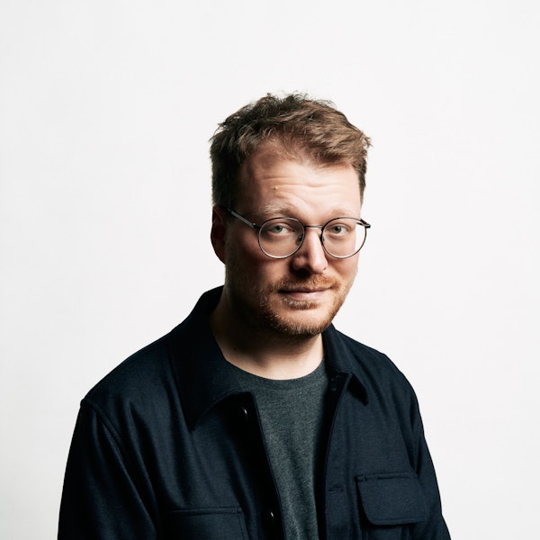 Stand-Up-Comedian Maxi Gstettenbauer steht vor einer weißen Wand. Er trägt ein blaues Hemd über einem grauen T-Shirt.