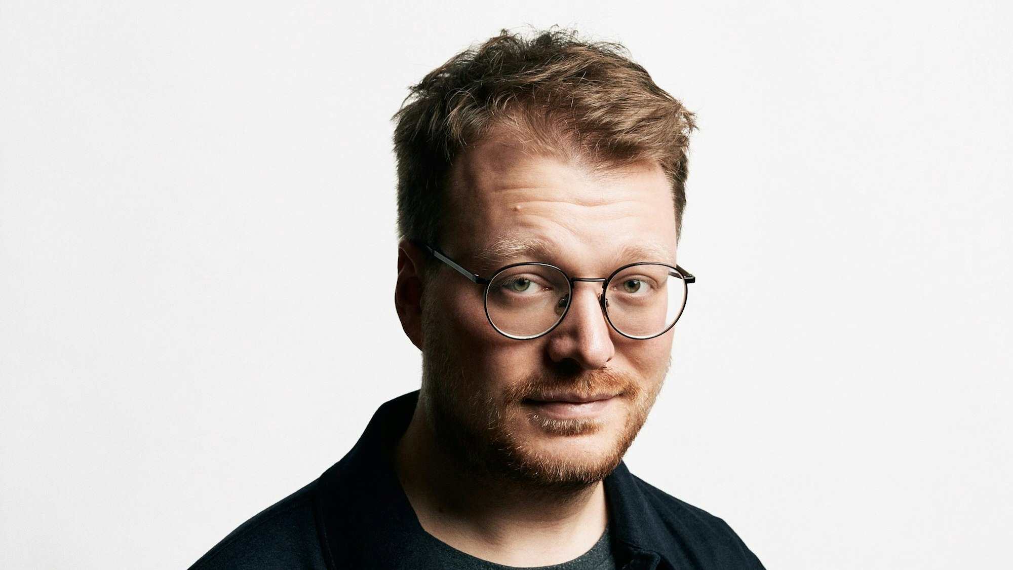 Stand-Up-Comedian Maxi Gstettenbauer steht vor einer weißen Wand. Er trägt ein blaues Hemd über einem grauen T-Shirt.