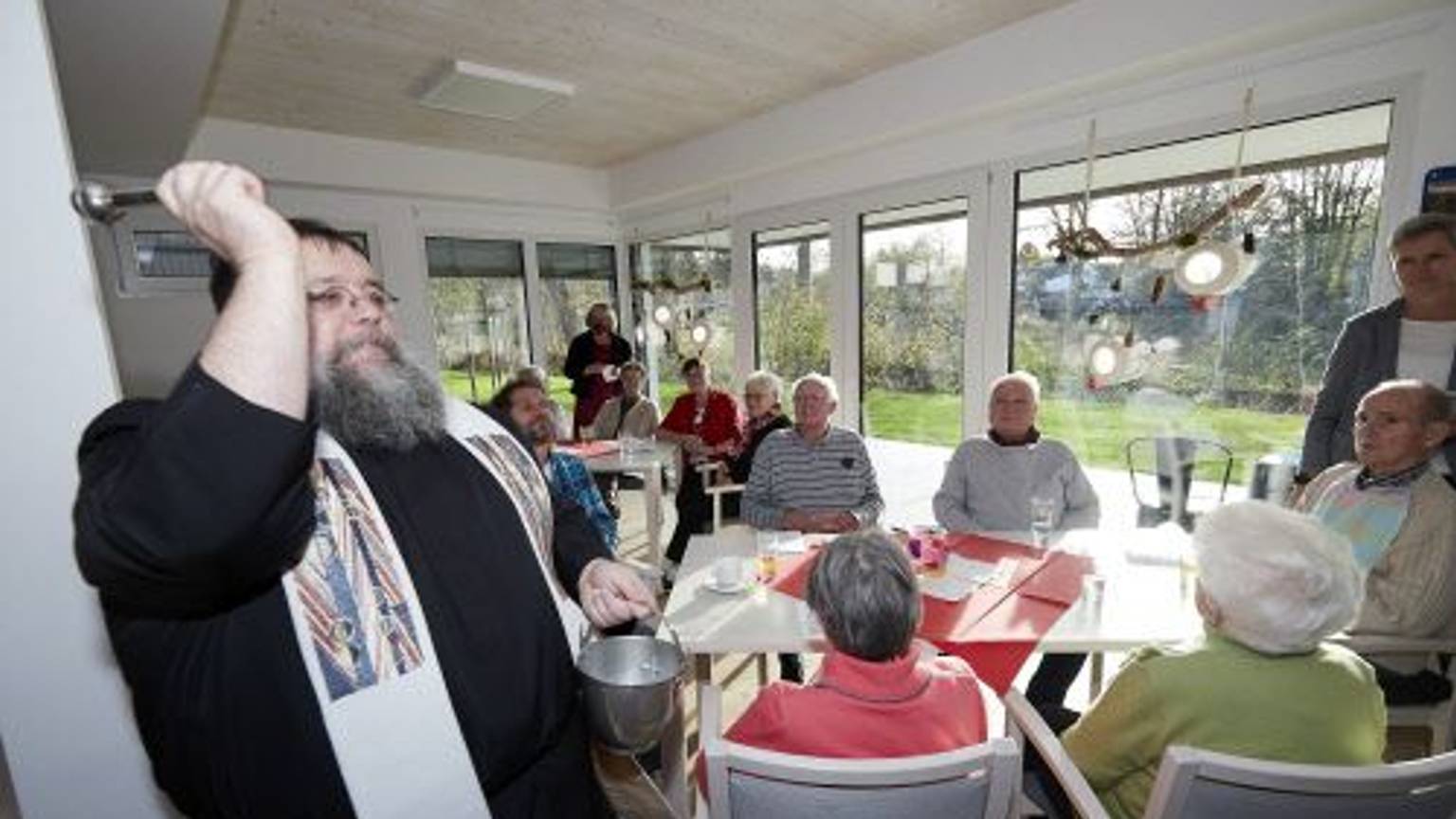 Pater Georg segnet einen Raum mit Weihwasser. Im Hintergrund sitzen Seniorinnen und Senioren an Tischen vor einer Fensterfront.