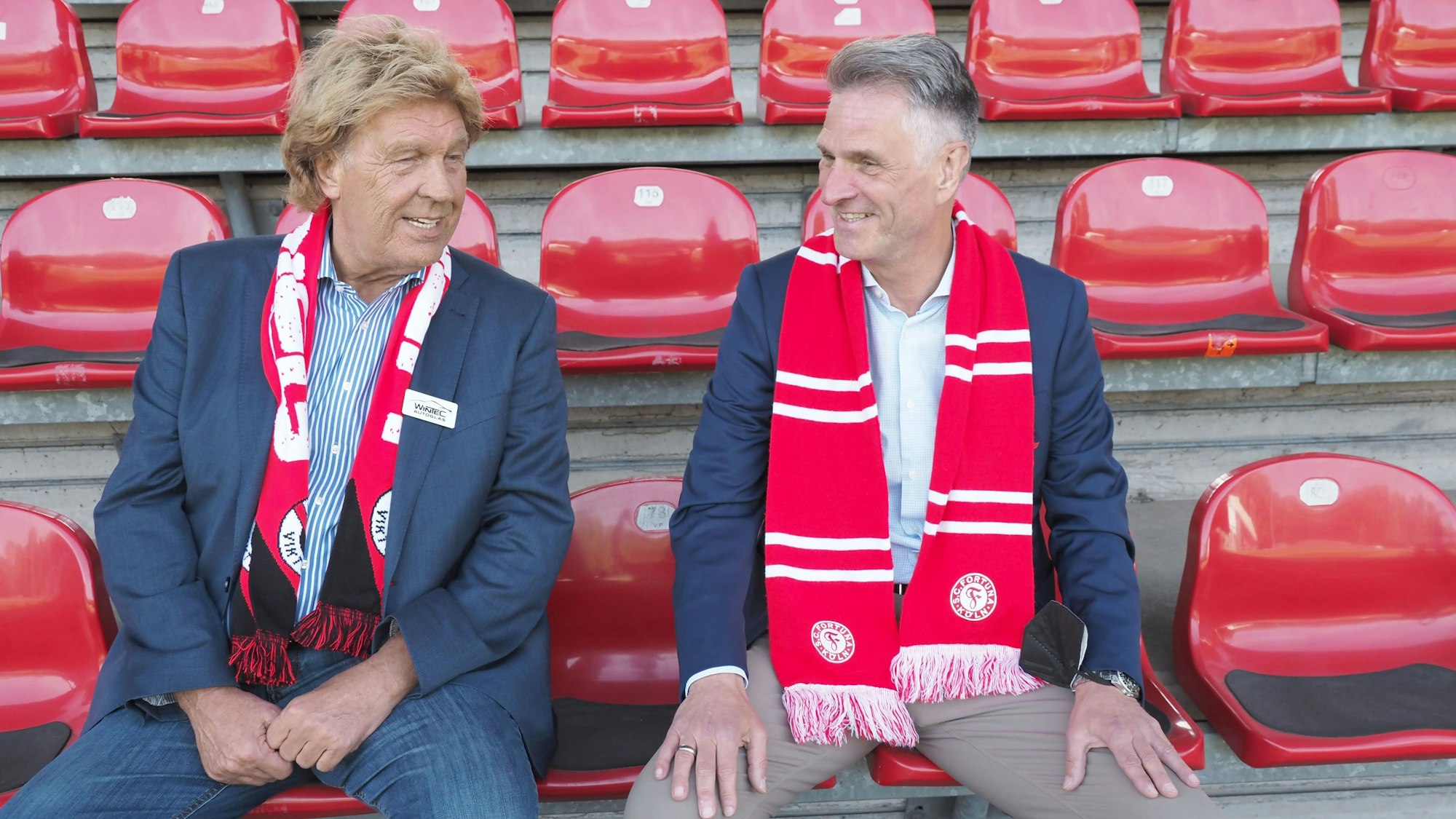 Zwei Männer sitzen auf der Tribüne eines Fußballstadions und unterhalten sich. Beide lächeln.