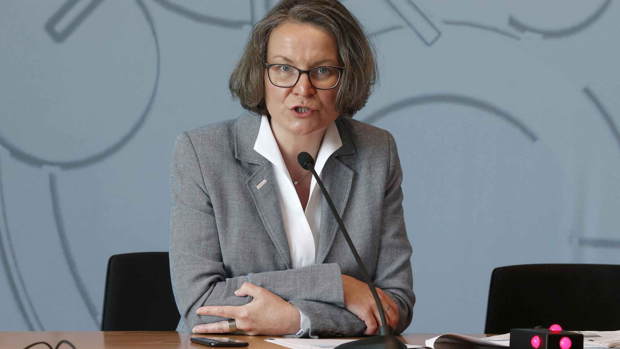 NRW-Ministerin Ina Scharrenbach (CDU) bei einer Pressekonferenz. Sie sitzt an einem Tisch, vor ihr ein schmales Mikrofon und hinter ihr eine graue Wand.
