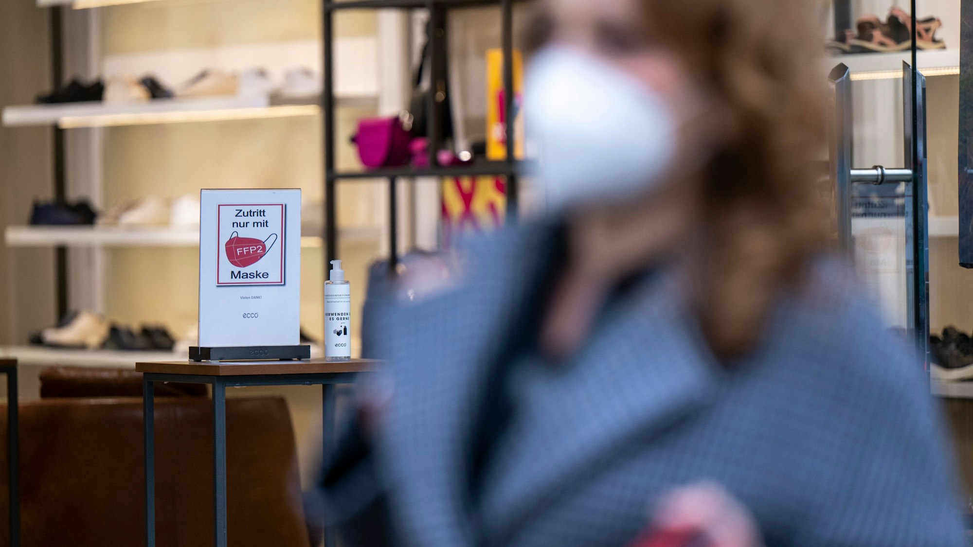 Ein Schild am Eingang eines Schuh-Geschäfts weist auf die Pflicht zum Tragen einer FFP2-Maske hin, während im Vordergrund eine Frau mit FFP2-Maske vorbeigeht.