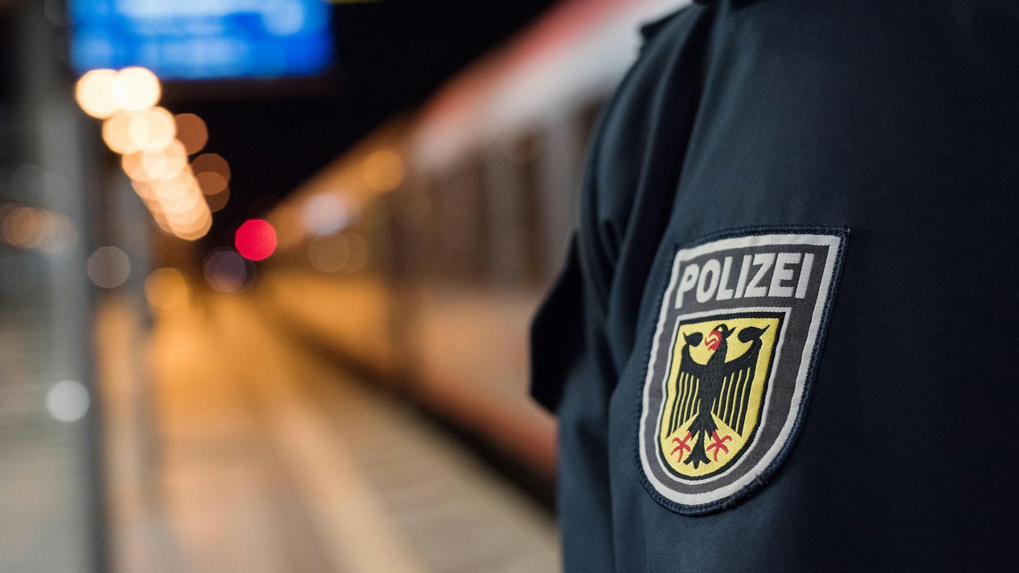 Auf der Jacke der Kölner Bundespolizei ist das Logo der Polizei. Im Hintergrund steht ein Zug auf den Schienen am Kölner Hauptbahnhof (Symbolbild)
