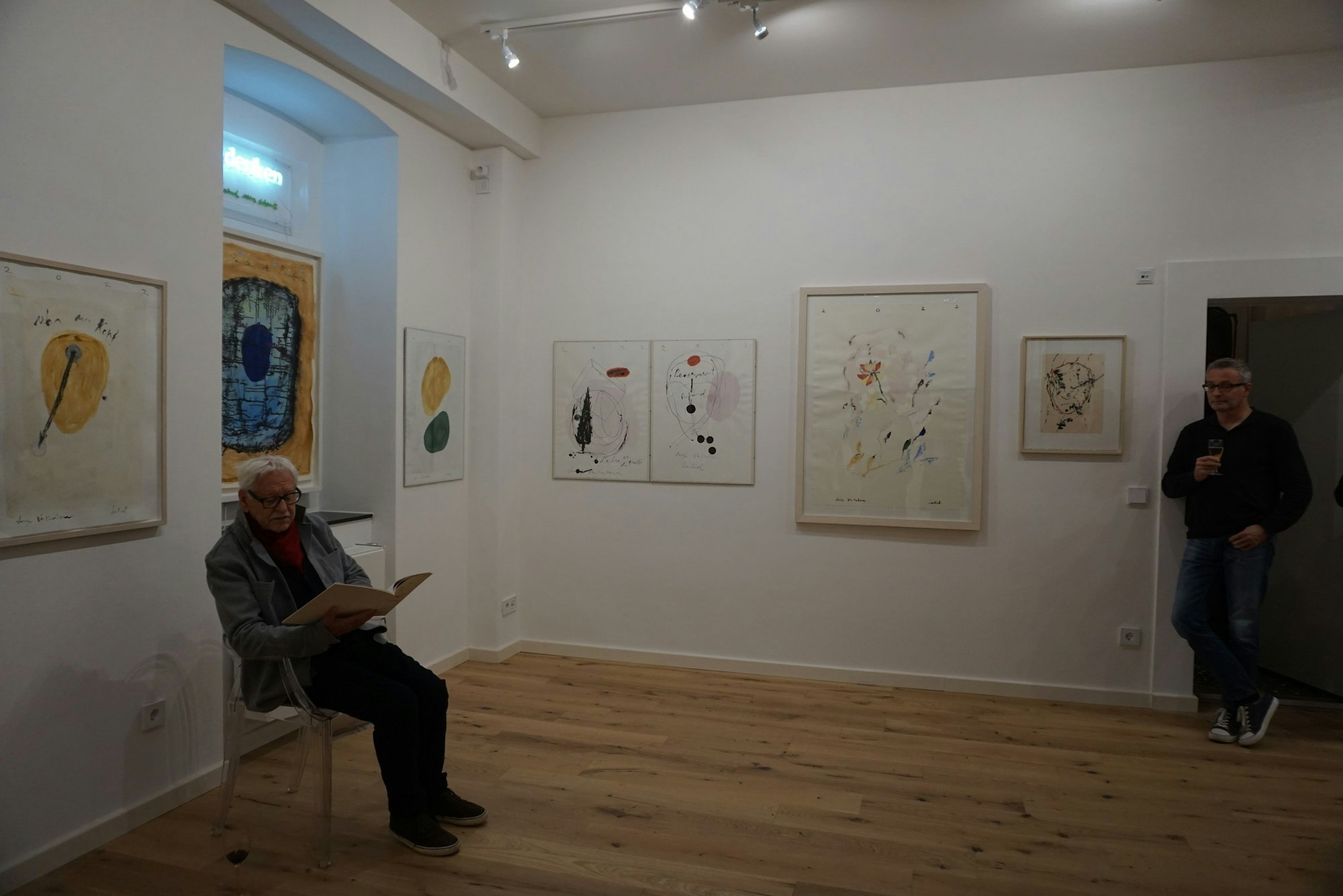 An den Wänden hängen mehrere Bilder. Ein Mann sitzt mit einem Buch auf einem Stuhl.