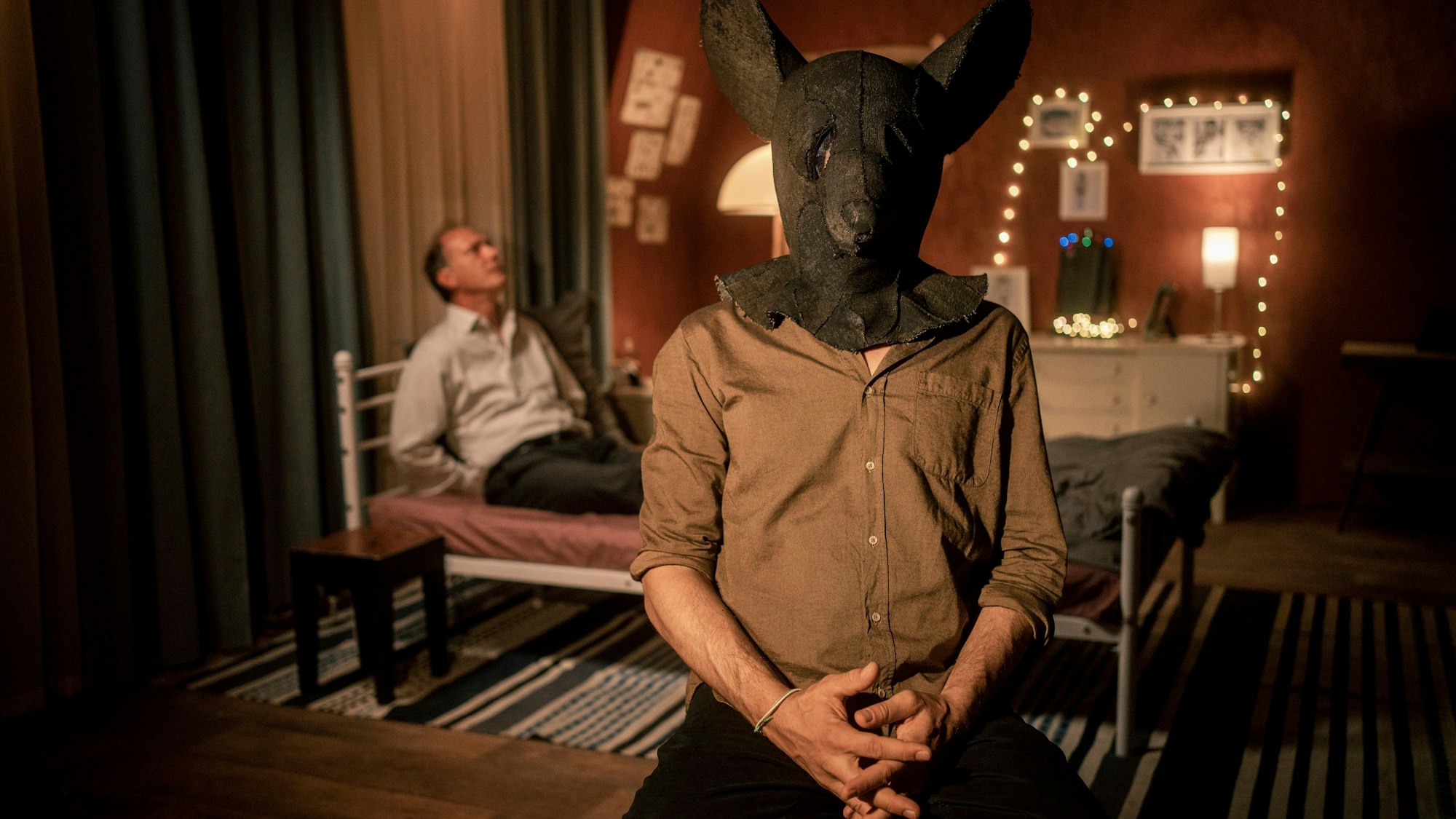 Der Entführer mit einer Mausmaske aus Folz nimmt ein Video auf. Er hat Peter Schnabel entführt, der hinter ihm an ein Bett gekettet ist.