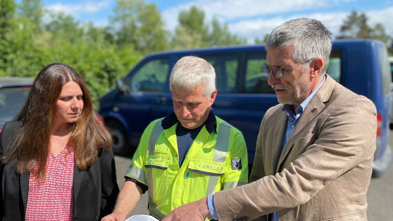 Eine Frau und zwei Männer stehen auf einem Parkplatz und schauen auf eine Karte.