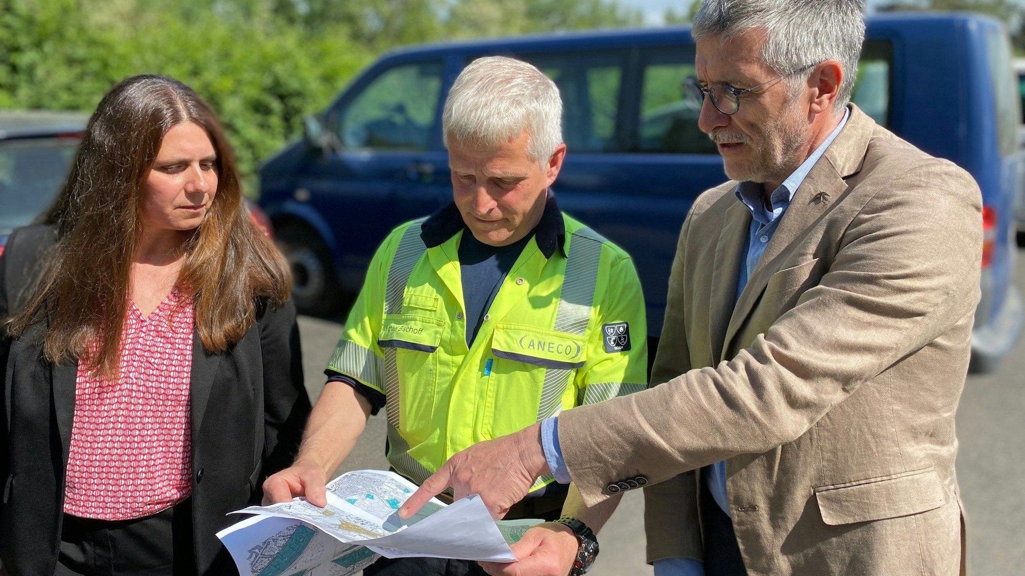 Eine Frau und zwei Männer stehen auf einem Parkplatz und schauen auf eine Karte.