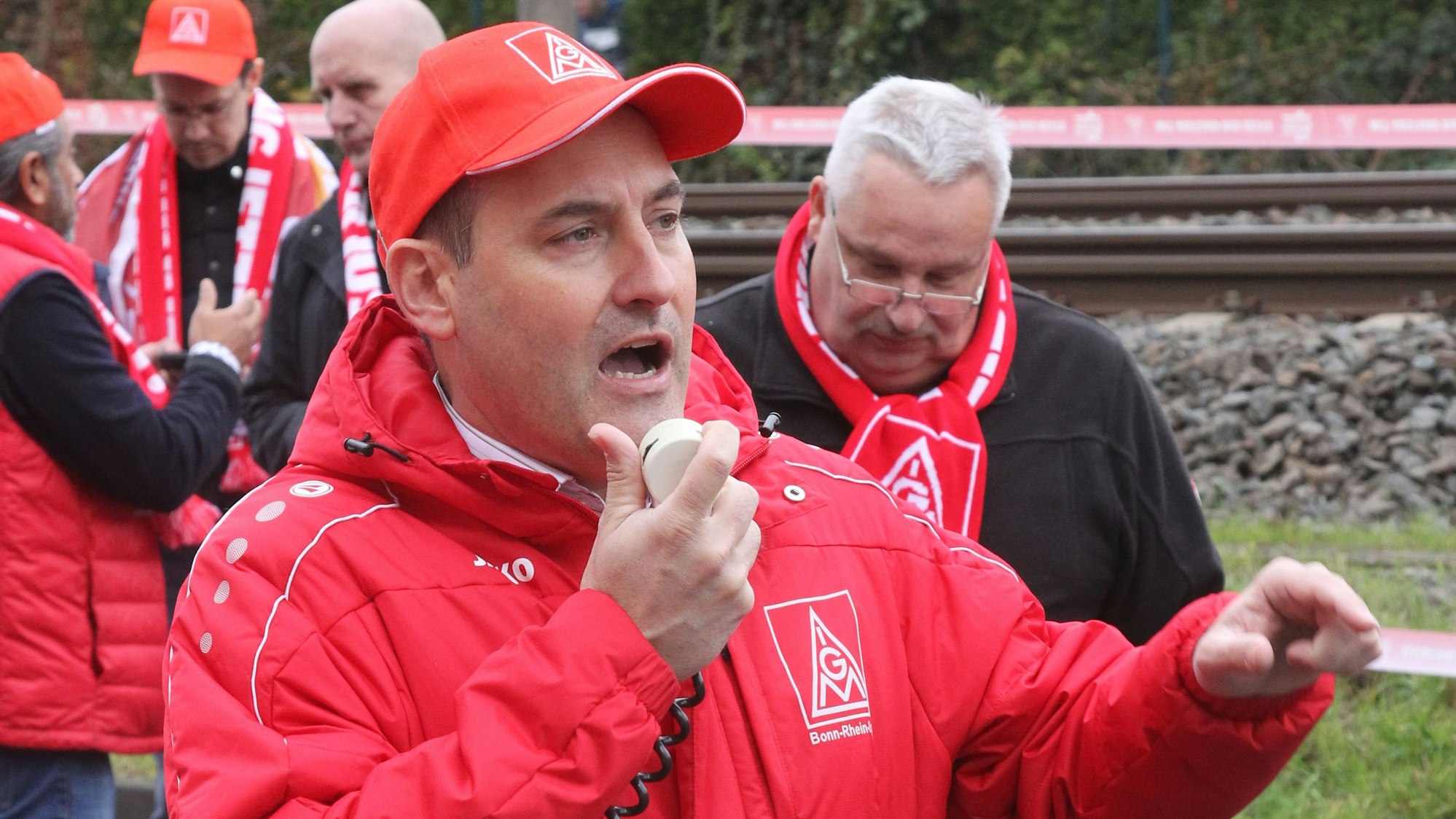 Michael Korsmeier in roter IG Metall Jacke und mit roter IG Metall Kappe auf dem Kopf spricht in ein Mikrofon.