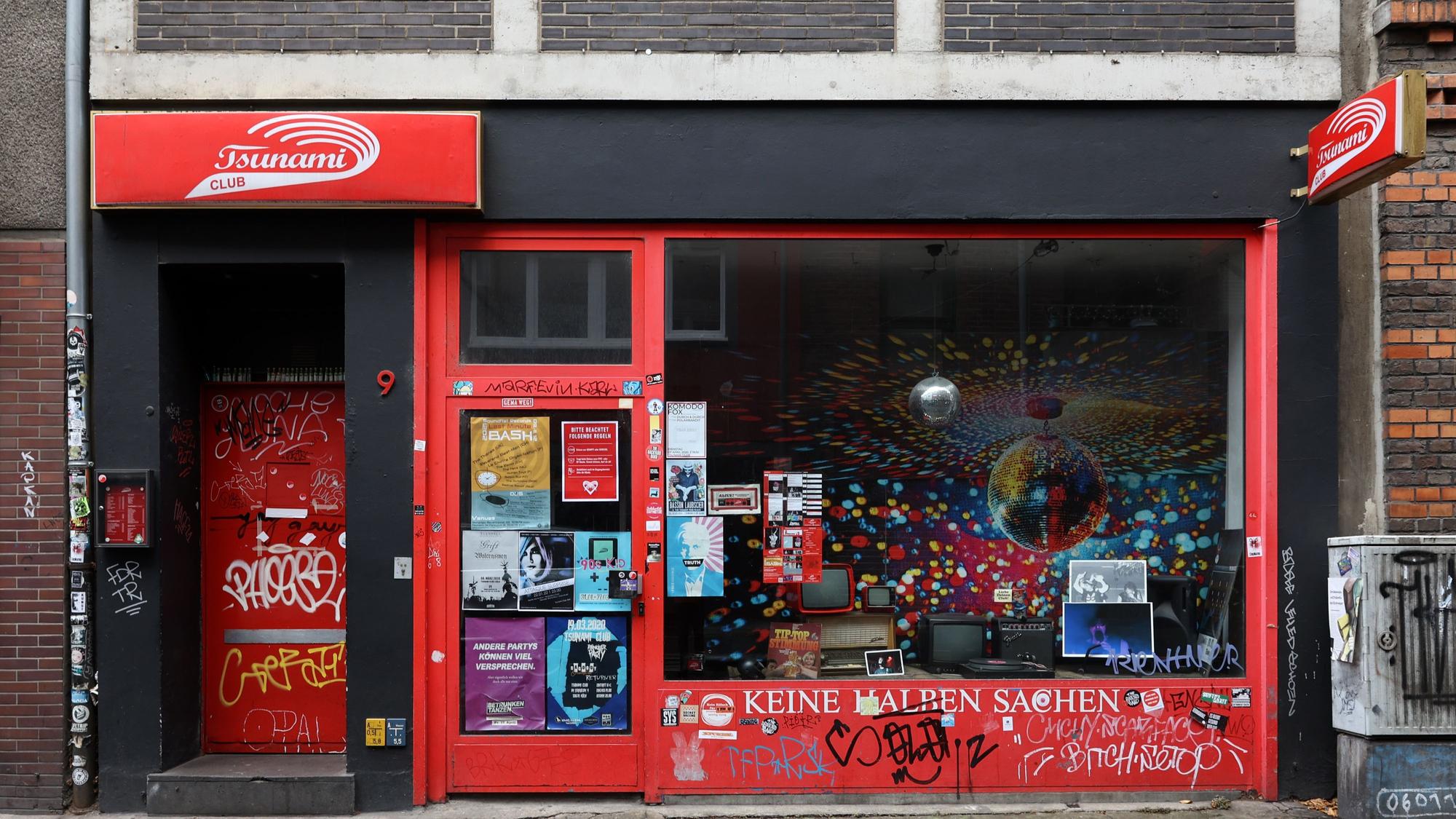 Die Fassade des Tsunami Clubs bei Tag, mit rotem Schriftzug, roter Tür und Schaufenster.