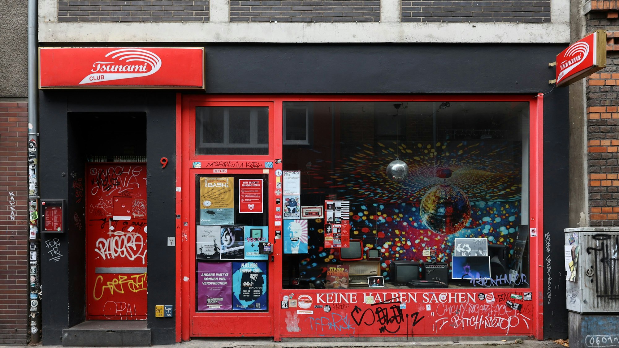 Die Fassade des Tsunami Clubs bei Tag, mit rotem Schriftzug, roter Tür und Schaufenster.
