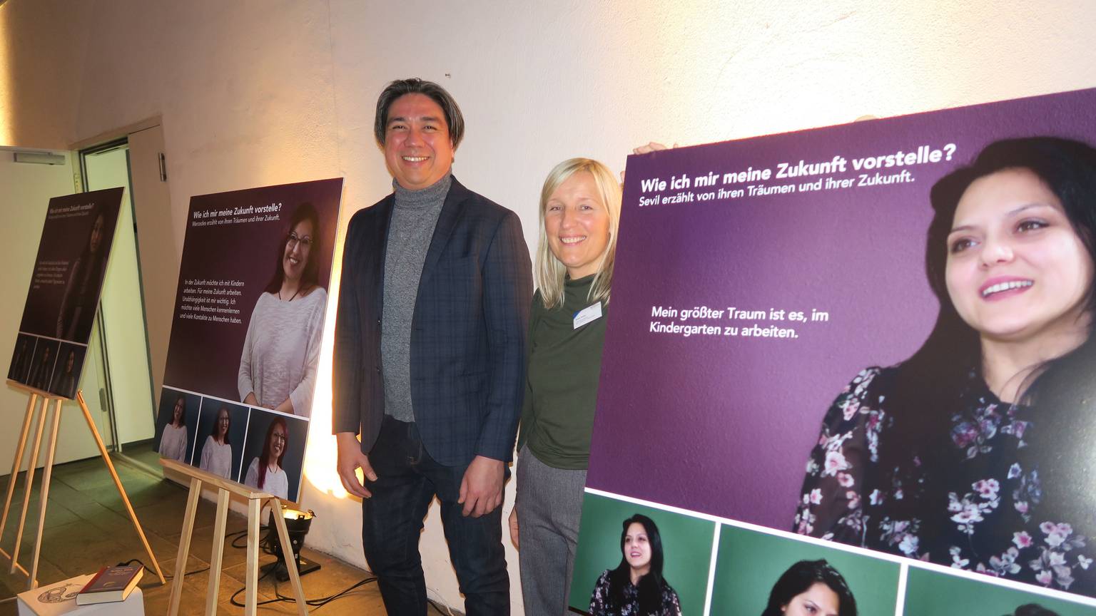Jhon Jimenez und Anna Drahs in der Ausstellung. Neben ihm sind Plakate aufgestellt, auf denen Bilder von jungen Frauen zu sehen sind. Neben den Frauen sind Zitate von diesen dargestellt.