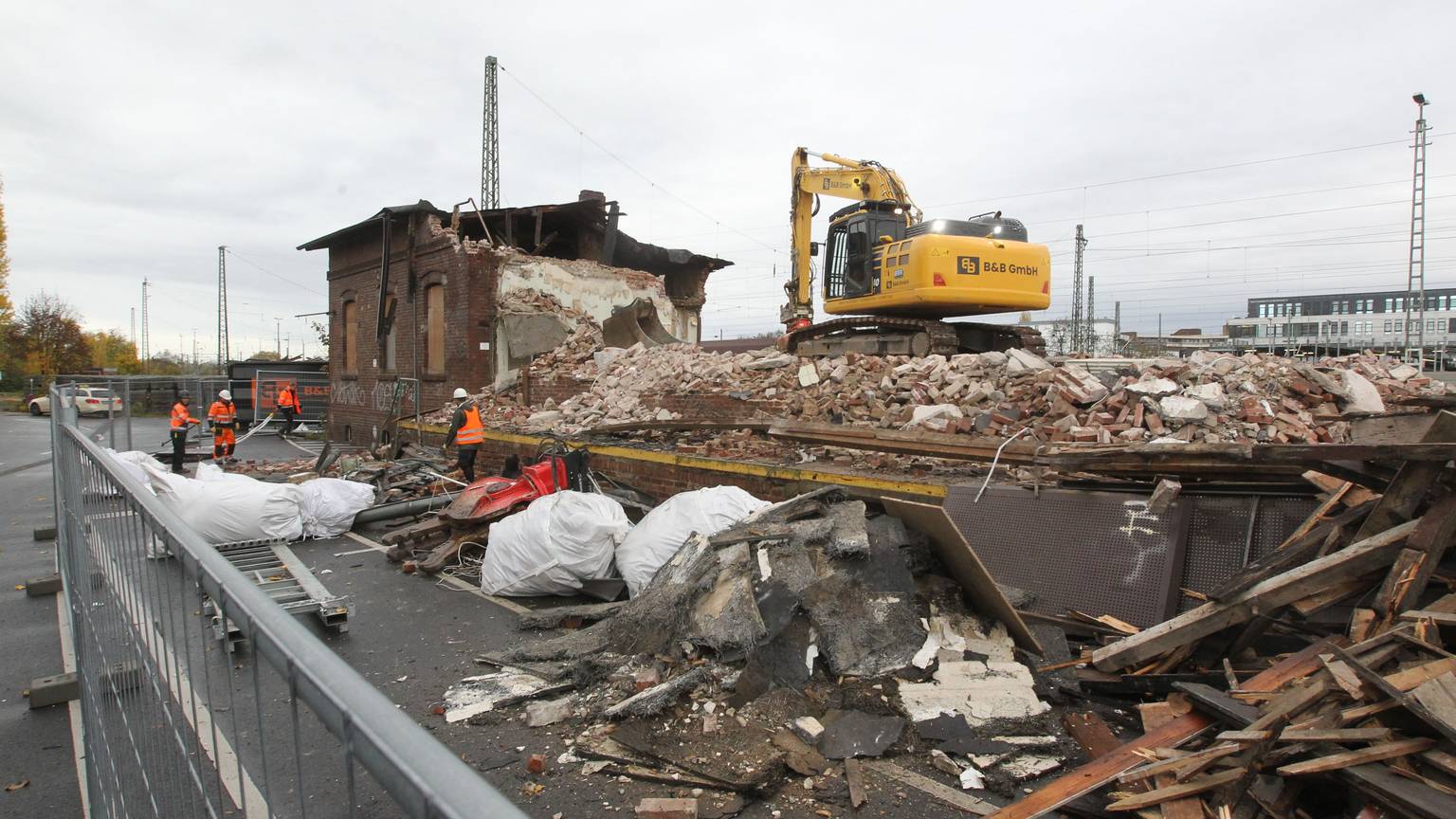 Ein gelber Bagger steht auf den Trümmern der Lagerhalle. Im Hintergrund ist noch ein Teil des Gebäudes aus Backsteinen zu sehen. Männer in orangen Sicherheitswesten arbeiten auf der Baustelle.
