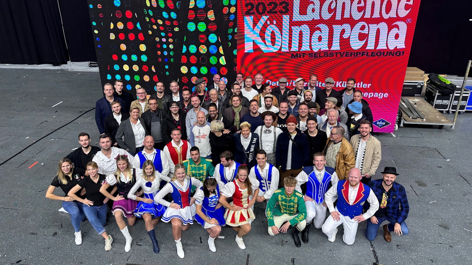 Rund 50 Mitwirkende der Lachenden Kölnarena 2023, Topbands des Kölner Karnevals und Tanzgruppenmitglieder, präsentieren sich vor einem digitalen Großplakat.