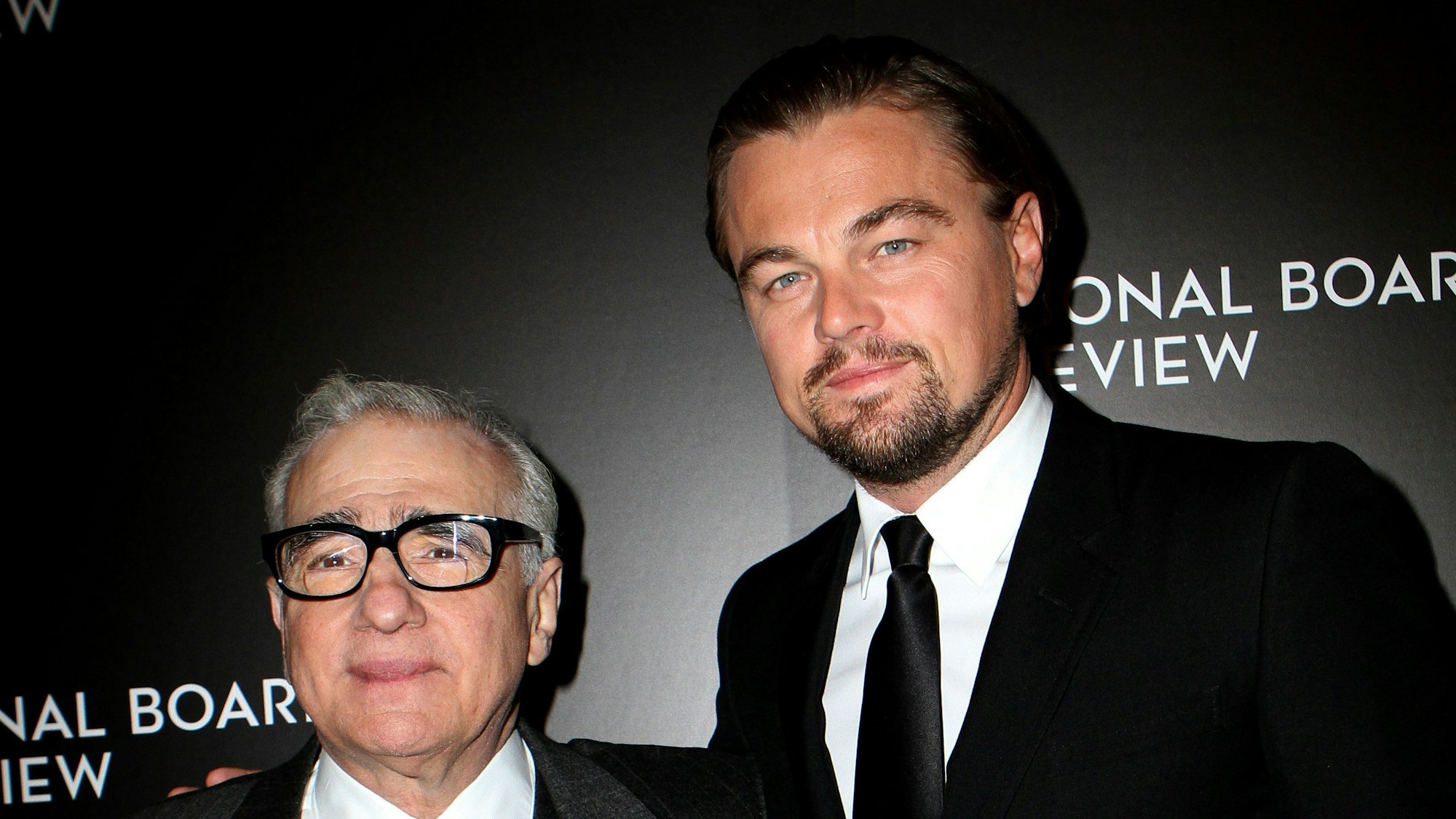 Leonardo DiCaprio und Martin Scorsese in schwarzen Anzügen. Sie stehen vor einer schwarzen Wand und lächeln.