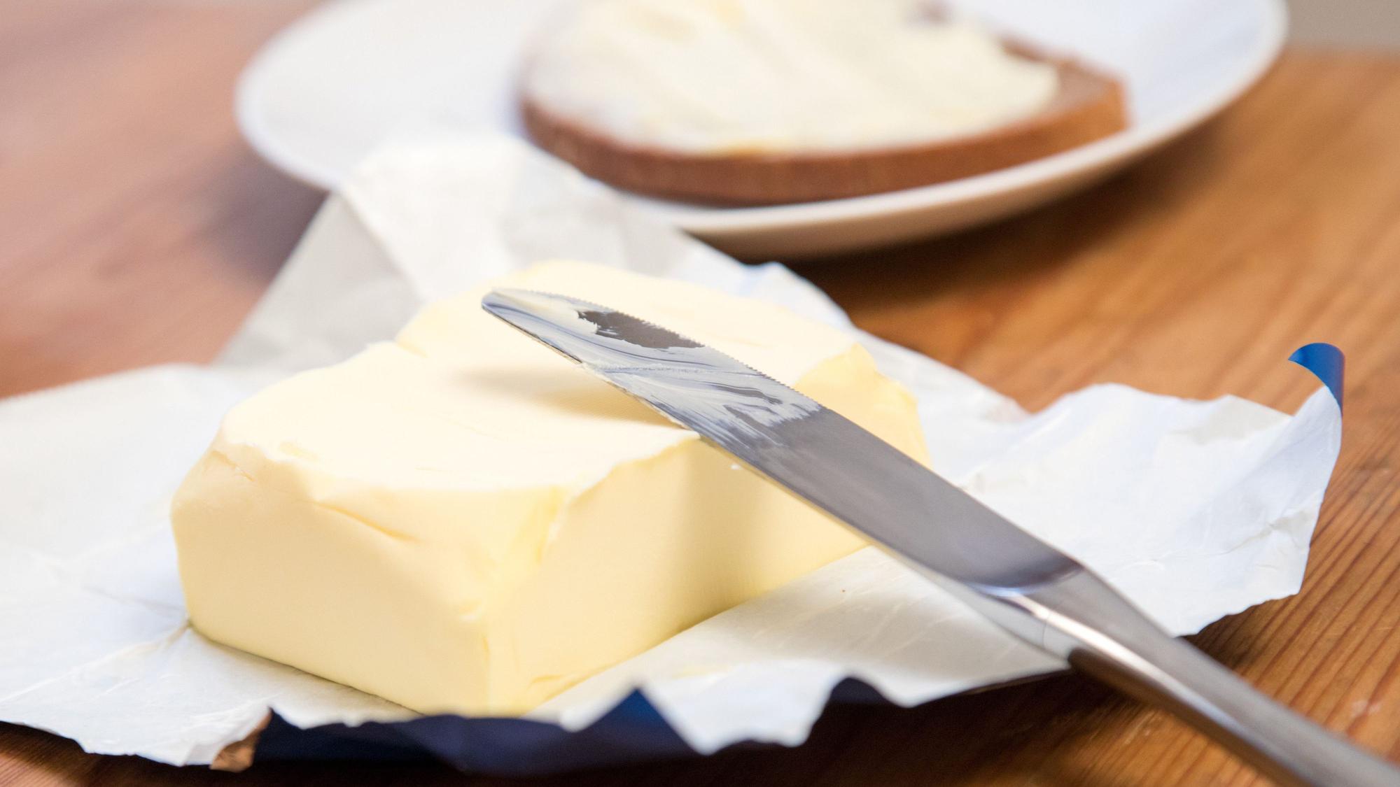 Ein Stück Butter und ein Messer liegen auf einem Tisch, im Hintergrund ist ein Butterbrot zu sehen.