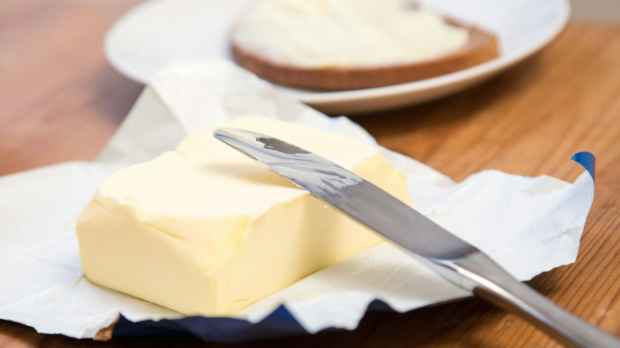 Ein Stück Butter und ein Messer liegen auf einem Tisch, im Hintergrund ist ein Butterbrot zu sehen.