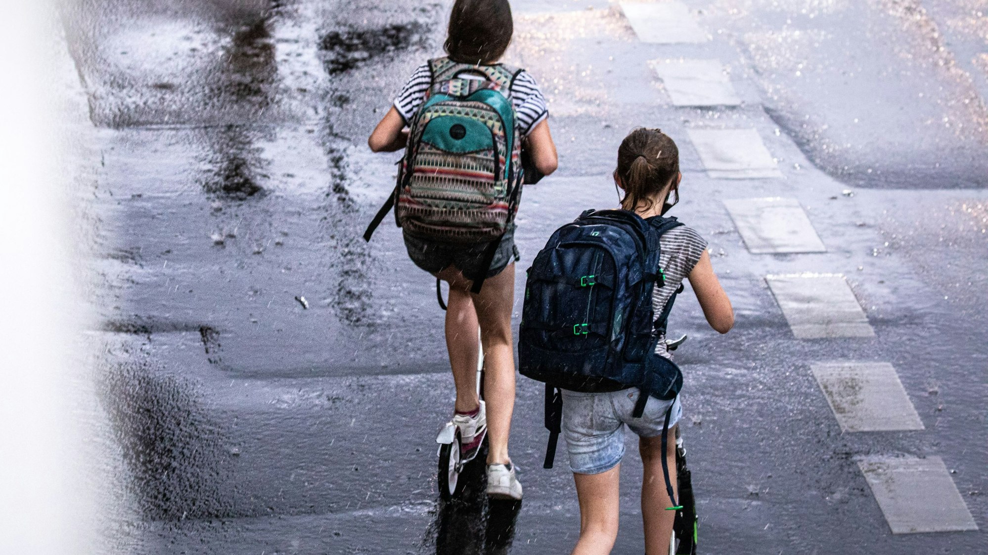 Zwei Schulkinder sind auf ihren Tretrollern unterwegs während eines Unwetters mit Gewitter und Starkregen im Ortsteil Sülz.