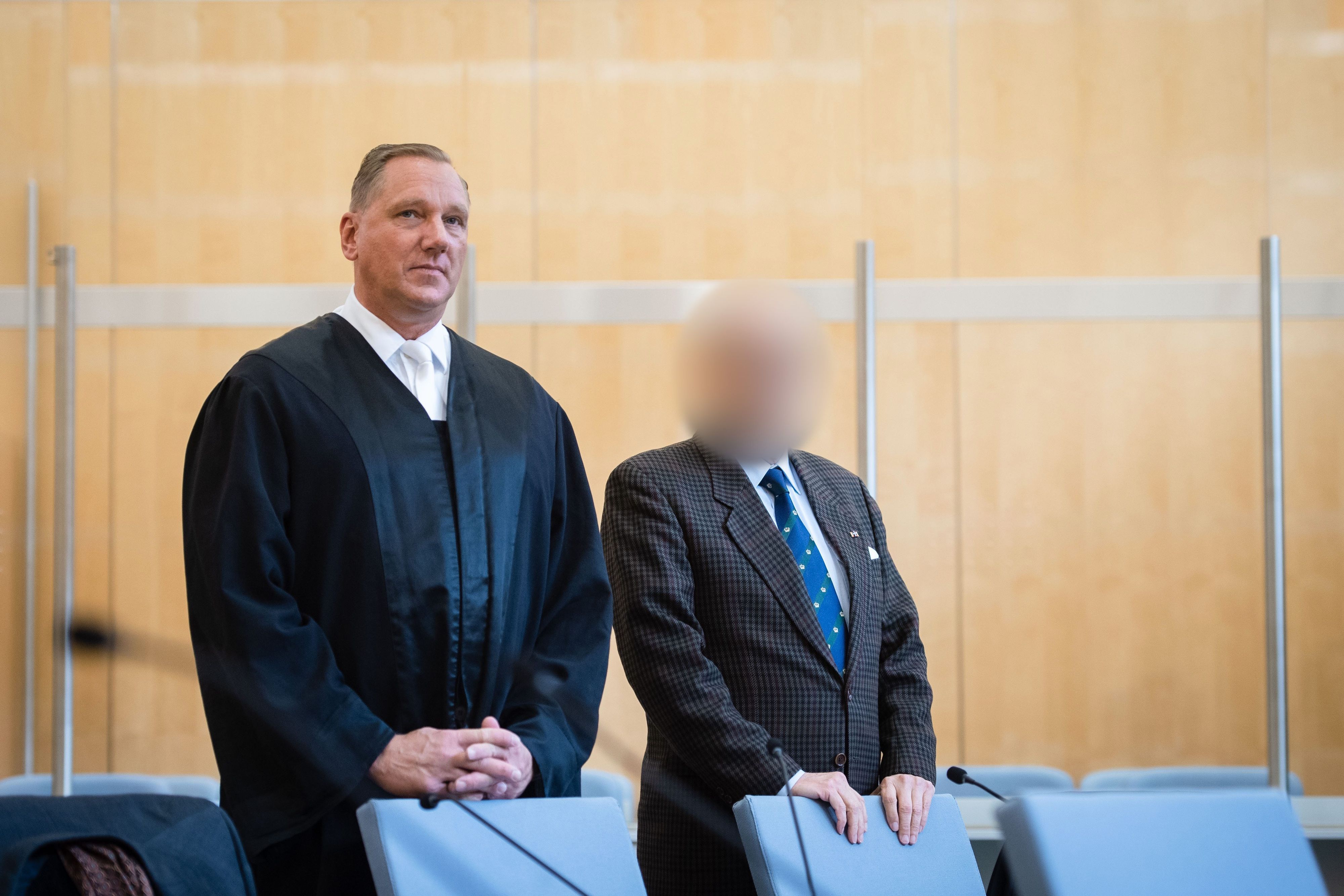 Blick in den Düsseldorfer Gerichtssaal, ein Angeklagter steht im Anzug hinter einem Stuhl, auf dem er lehnt, sein Gesicht ist nicht zu erkennen. Sein Verteidiger steht neben ihm.