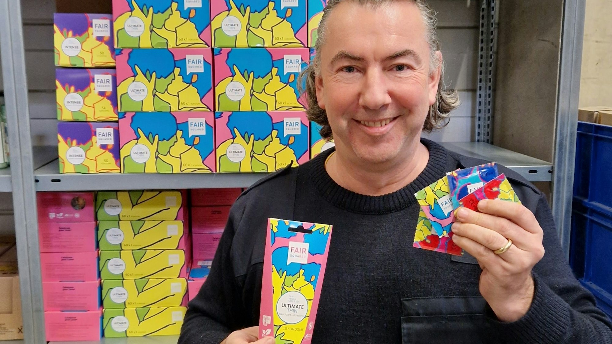 Oliver Gothe steht vor einem Regal und hält mehrere Packungen seiner Öko-Kondome in der Hand.