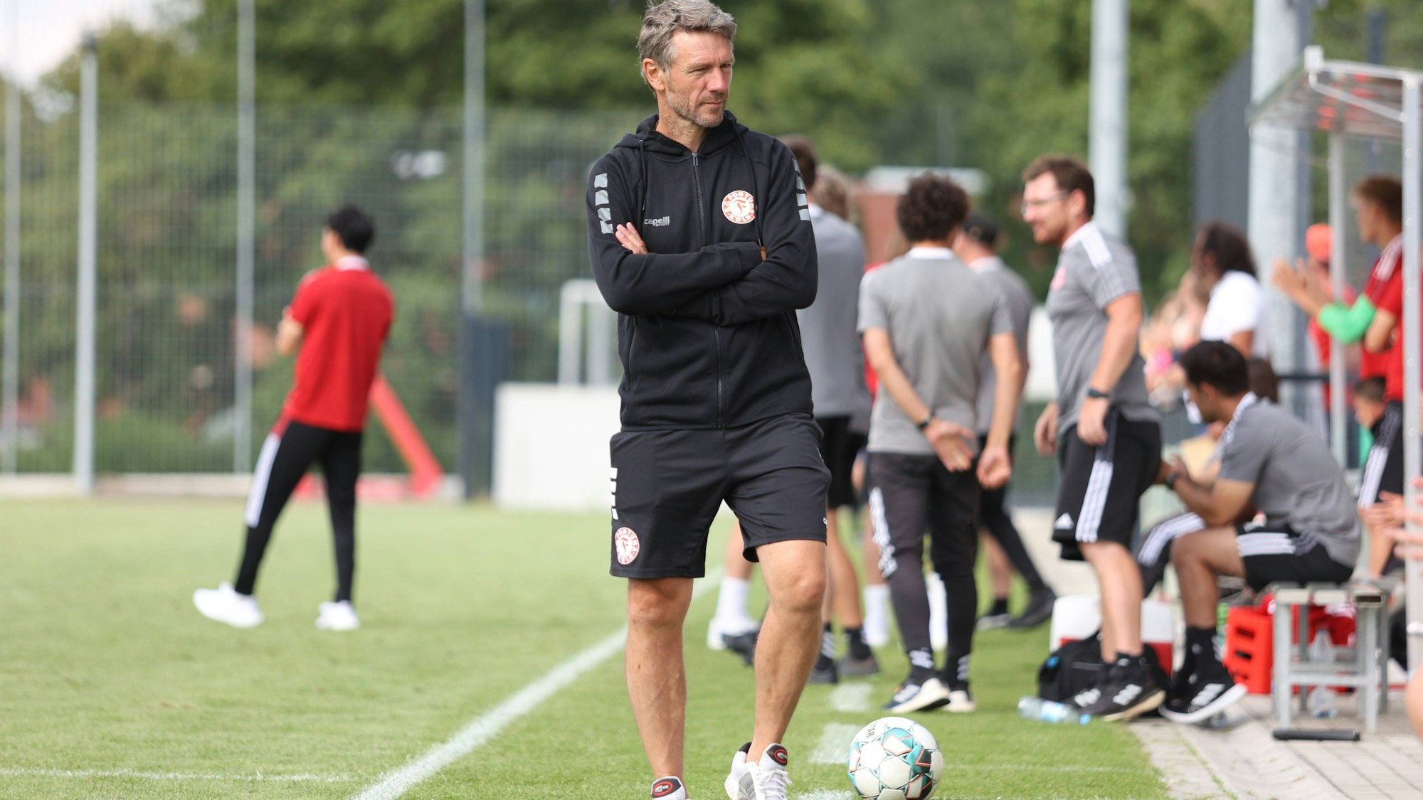 Ein Fußballtrainer geht mit verschränkten Armen am Spielfeldrand entlang und richtet den Blick auf die Auswechselbank.
