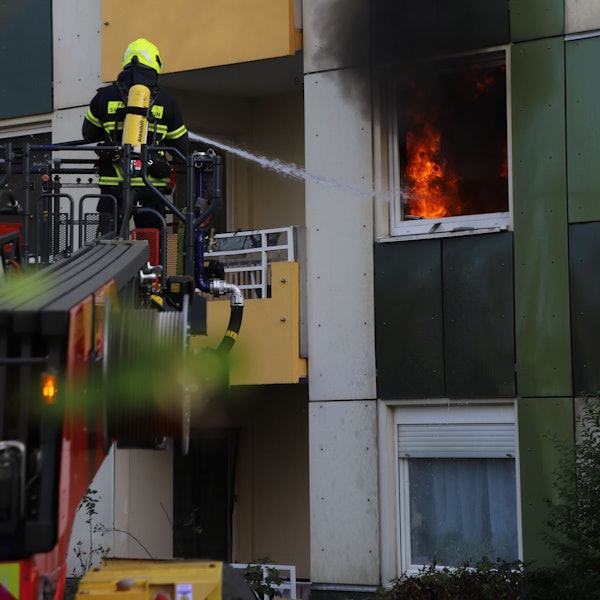Ein Feuerwehrmann steht auf einer Drehleiter und löscht mit einem Schlauch das Feuer hinter einem Fenster. Es steigt schwarzer Rauch auf.