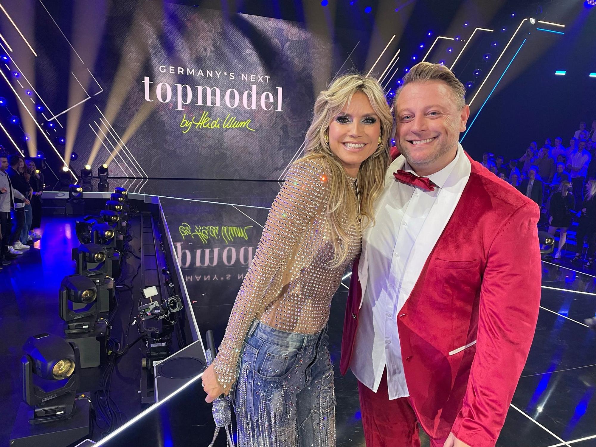 Christian Oberfuchshuber steht neben Heidi Klum auf der Bühne vom Finale der Castingshow „Germanys Next Topmodel“. Die beiden legen den Arm umeinander und lächeln in die Kamera.