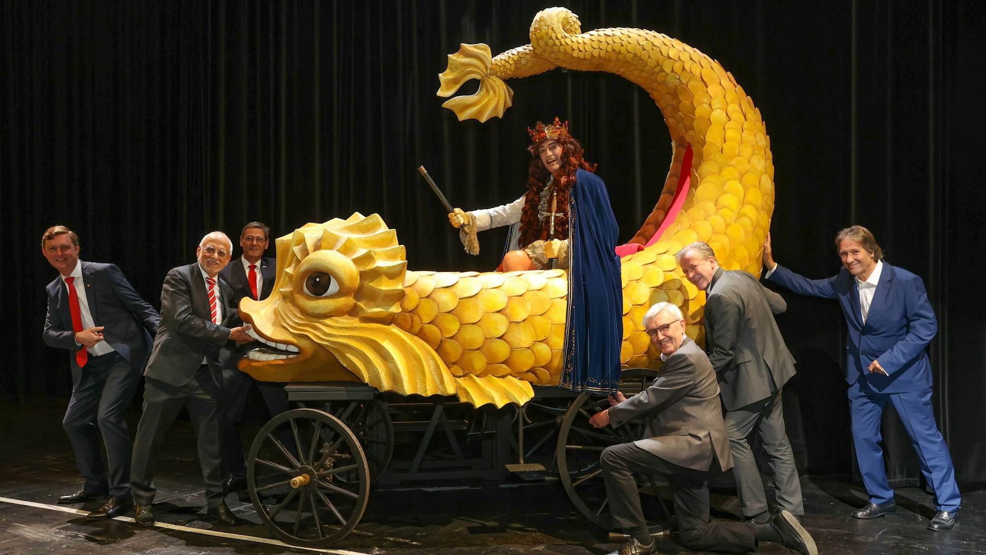 Übergabe des historischen Festwagens an den „Held Carneval“ (Stefan F. Robert). Sechs Männer stehen um einen kleineren Karnevalswagen herum. Auf dem Wagen sitzt der „Held Carneval“ auf einem gelben Wasser-Fabelwesen.