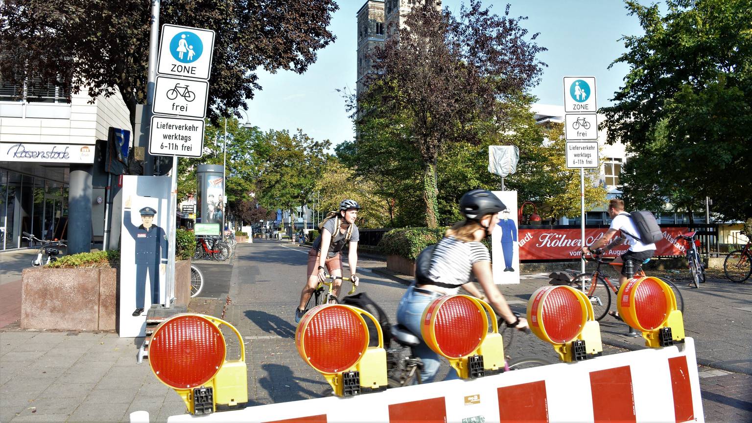 Schilder und Baken markieren das Durchfahrtsverbot für Autos auf der Deutzer Freiheit. Im Vordergrund sieht man Radfahrer, im Hintergrund steht St. Heribert.