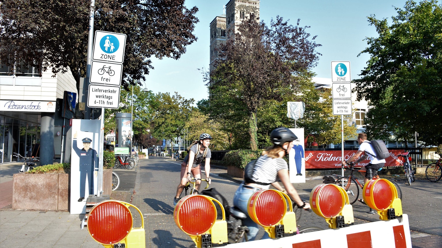 Schilder und Baken markieren das Durchfahrtsverbot für Autos auf der Deutzer Freiheit. Im Vordergrund sieht man Radfahrer, im Hintergrund steht St. Heribert.