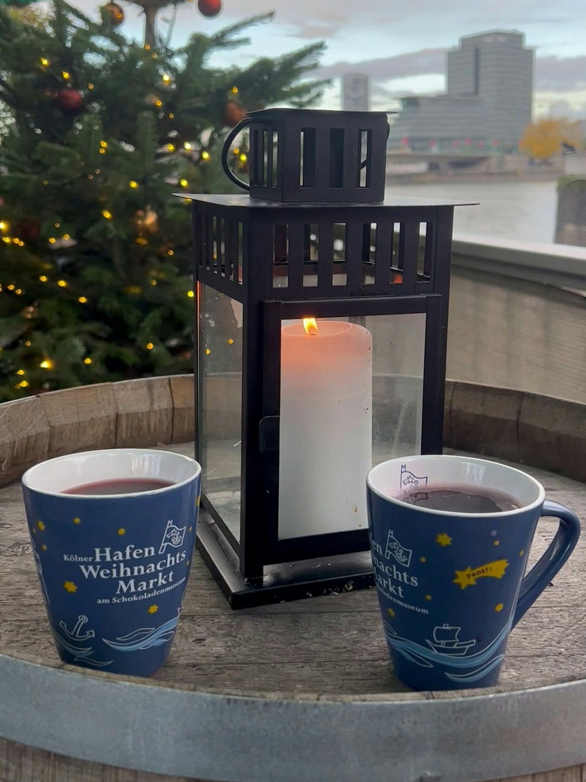 Zwei blaue Tassen vor einer Laterne mit Kerze auf einem Holzfass, das als Tisch dient, im Hintergrund der Rhein. November 2022.