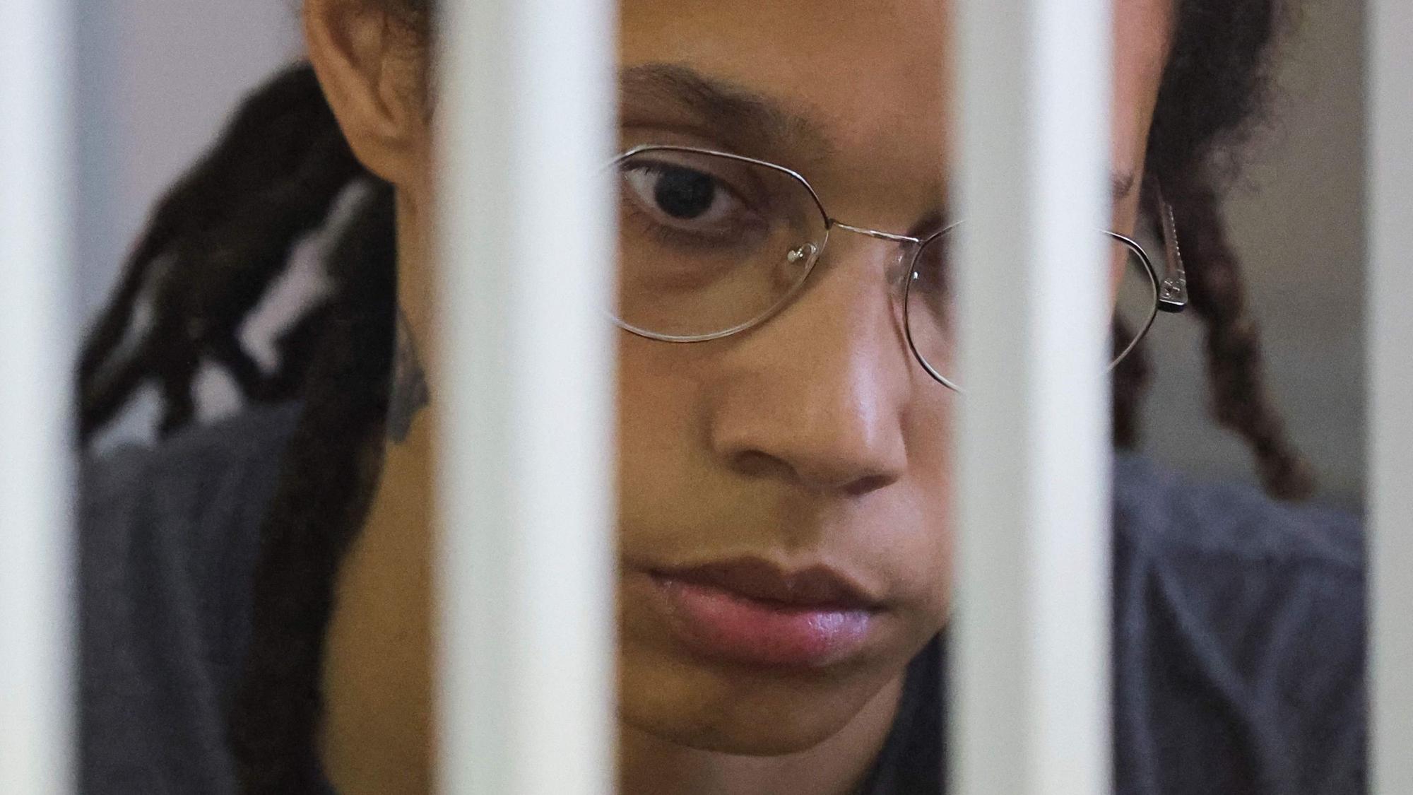 Brittney Griner Gesicht ist zu sehen, sie guckt traurig nach unten und wirkt abwesend. Vor ihrem Gesicht sieht man unscharf Gitterstäbe.