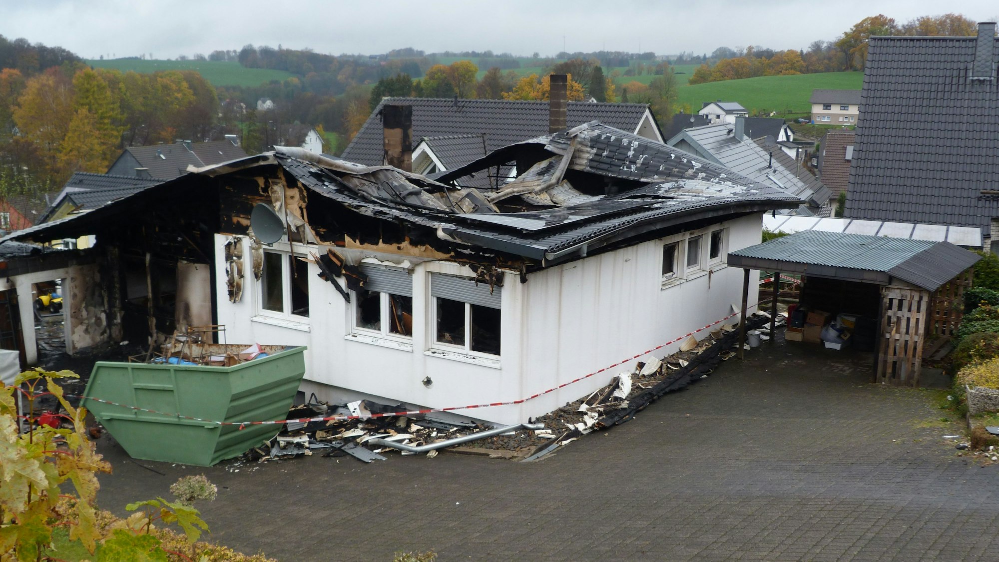 Das Dach eines Einfamilienhauses ist von einem Feuer vernichtet. Vor dem Haus steht ein Schuttcontainer.