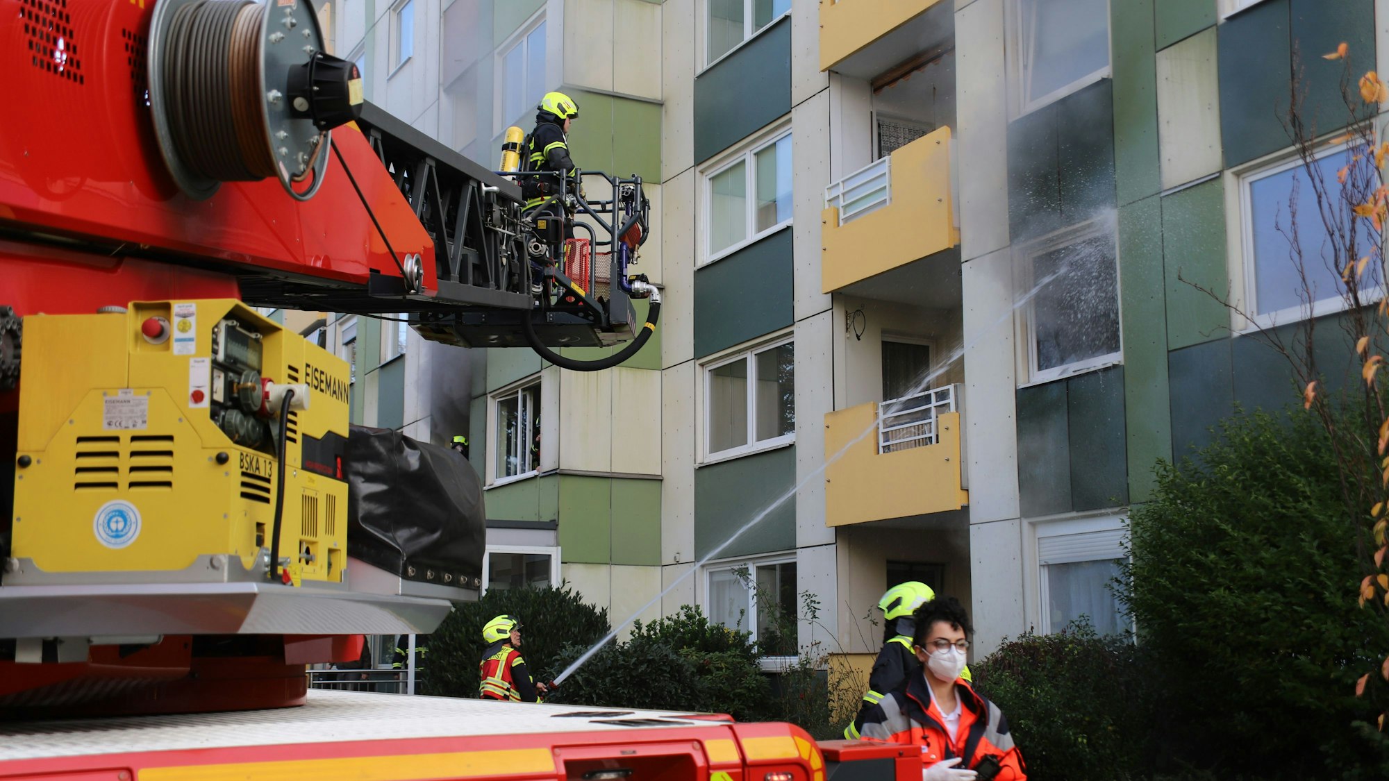 Das Feuerwehrauto mit Drehleiter steht vor einem Hochhaus. Ein Feuerwehrmann löscht mit einem Wasserstrahl vom Boden aus das Feuer.