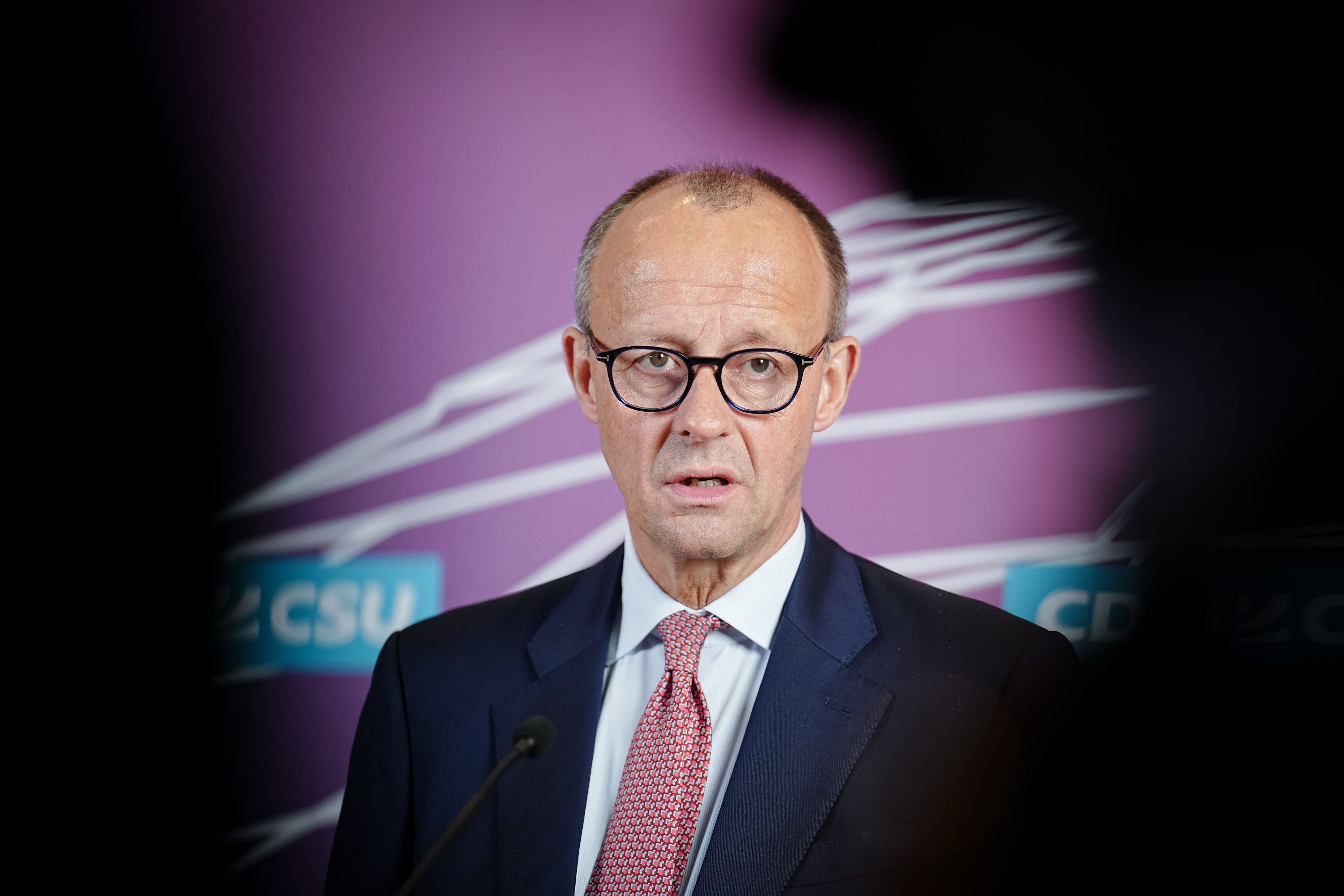 Merz steht vor einer Wand mit Unionslogo, redet und guckt in die Kamera. Am Rand des Bildes sieht man schwarze Schatten.