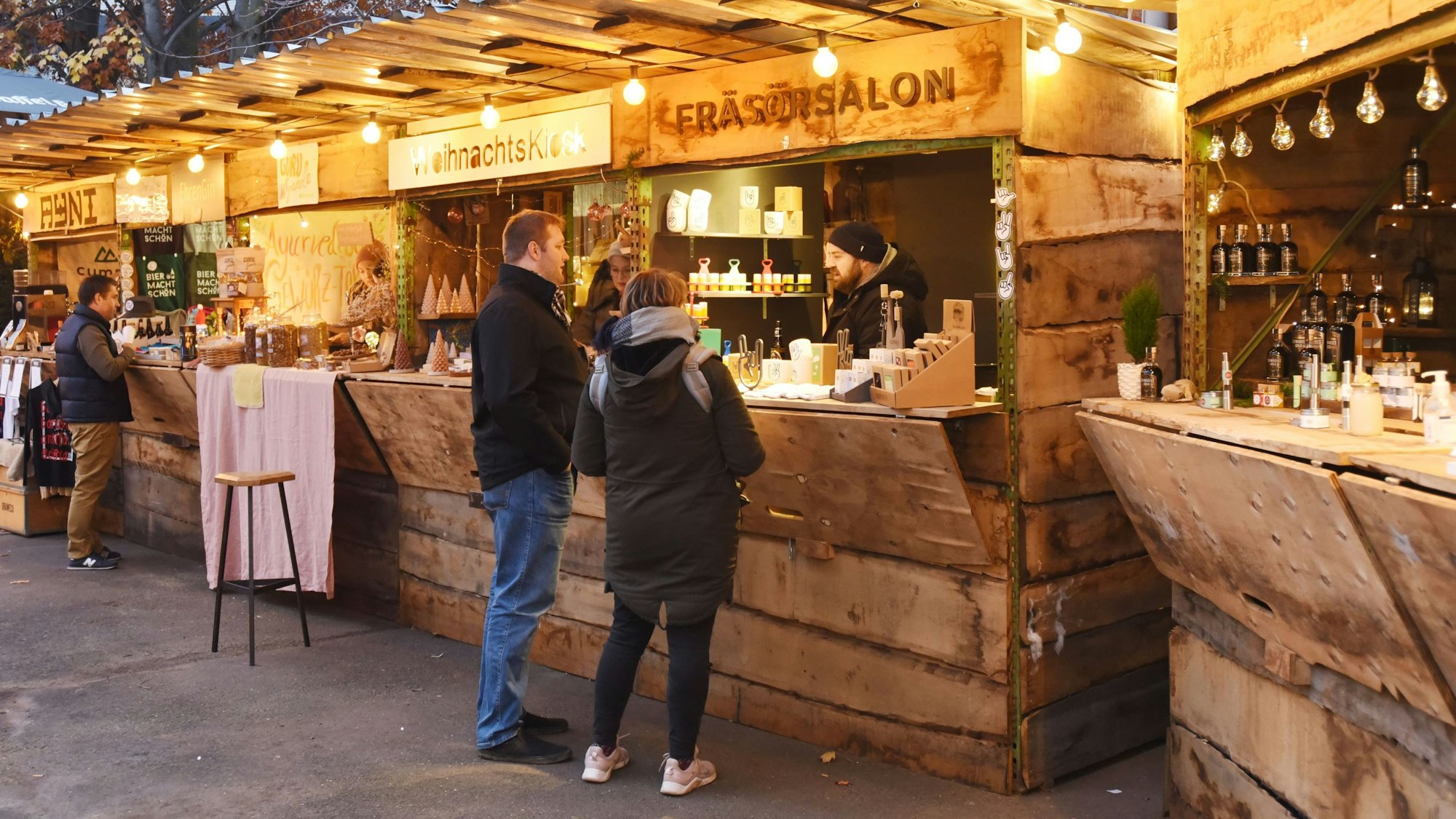 ein Mann und eine Frau stehen vor einer von mehreren Holzbuden, die von einer Lichterkette beleuchtet werden