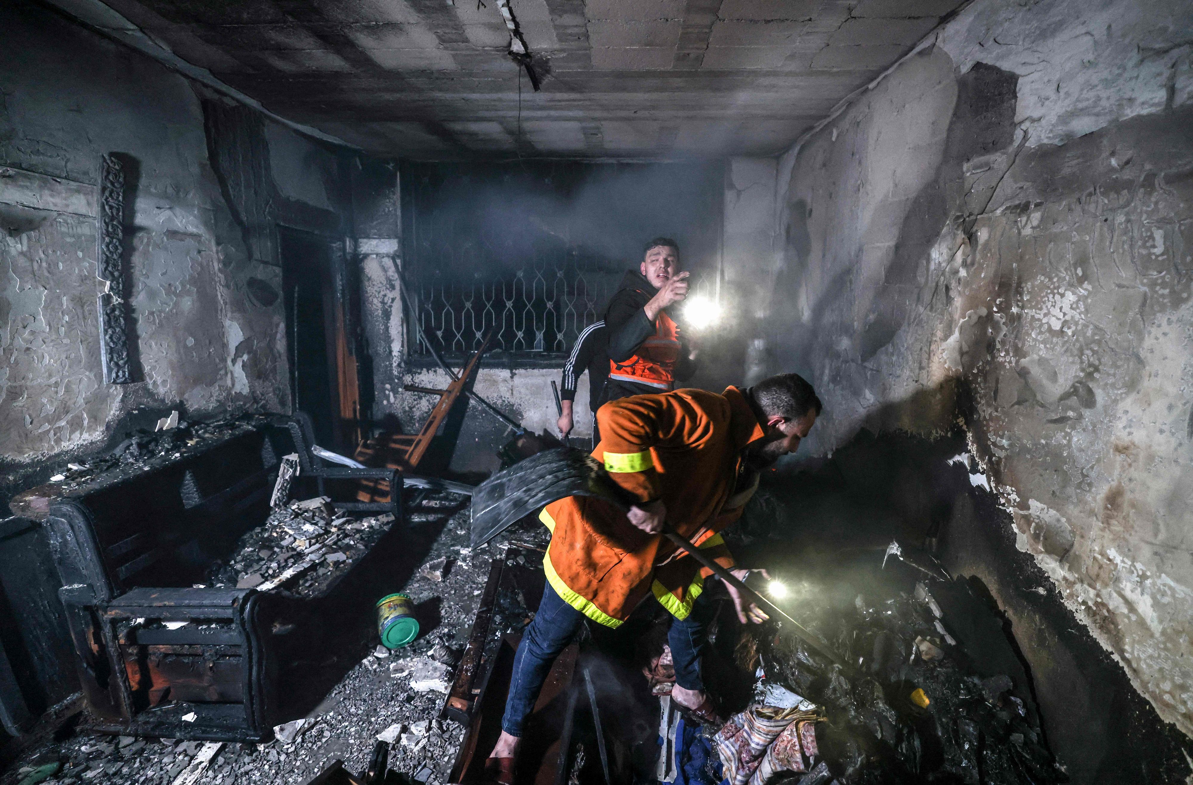 Man sieht zwei Männer in orangen Jacken, die mit Taschenlampen Wände und Boden absuchen. Das Bild zeigt einen Raum, in dem es gebrannt hat, links steht ein verkohltes Sofa, die Wände sind kahl, das Fenster an der hinteren Wand ist schwarz. In der Luft ist Staub oder Rausch.