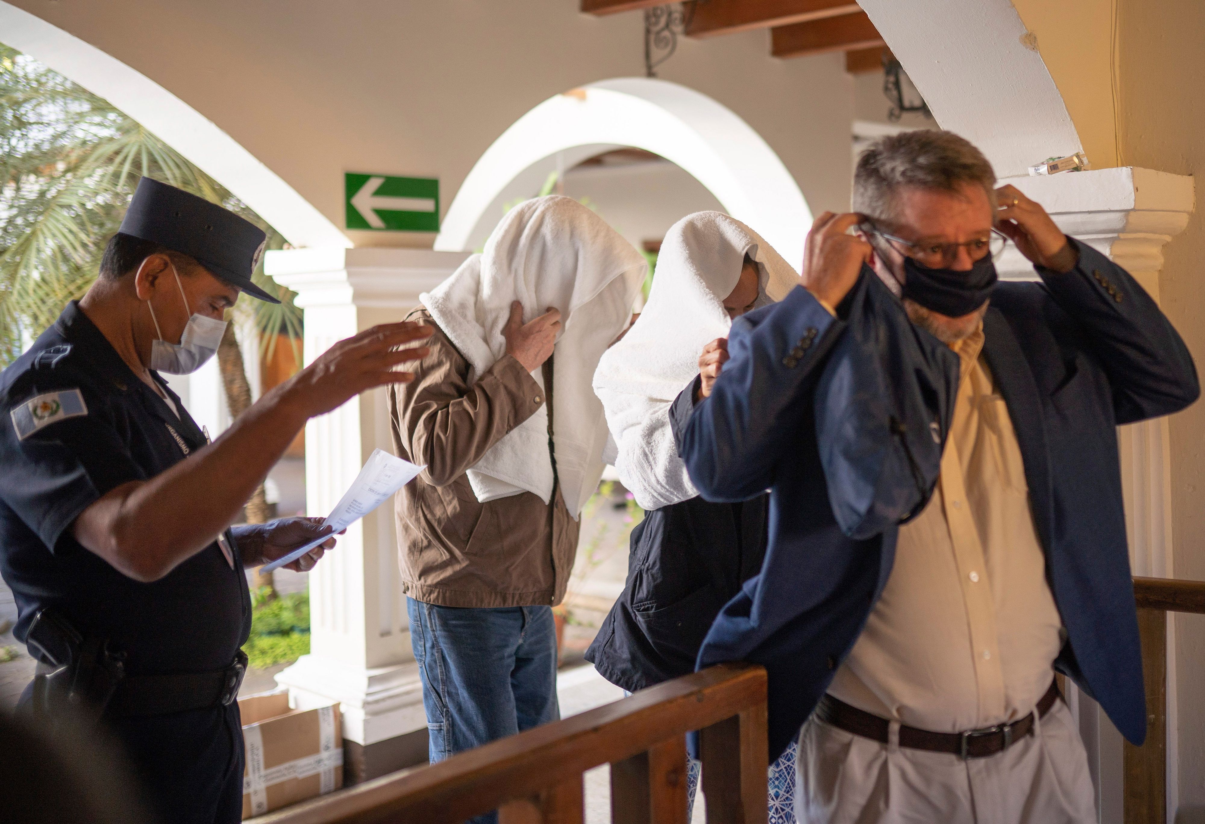 Zu sehen sind zwei Personen im Hintergrund, beide mit Handtüchern über dem Kopf, sie folgen einem Mann, der sich eine Maske anzieht. Links von ihnen steht ein Polizist, ebenfalls mit Maske, welcher ihnen den Weg weist und auf ein Blatt Papier schaut. Die Personen befinden sich in einem Gebäude und passieren eine Art Tor.