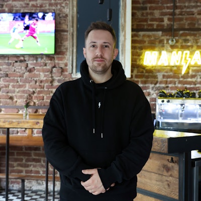 Nicolas Gottschalk, der Wirt der Fußballkneipe Brüsseler dem Brüsseler Platz, steht im Gastraum vor der Theke mit verschränkten Händen. An der Wand hängt ein Bildschirm, auf dem ein Fußballspiel gezeigt wird.