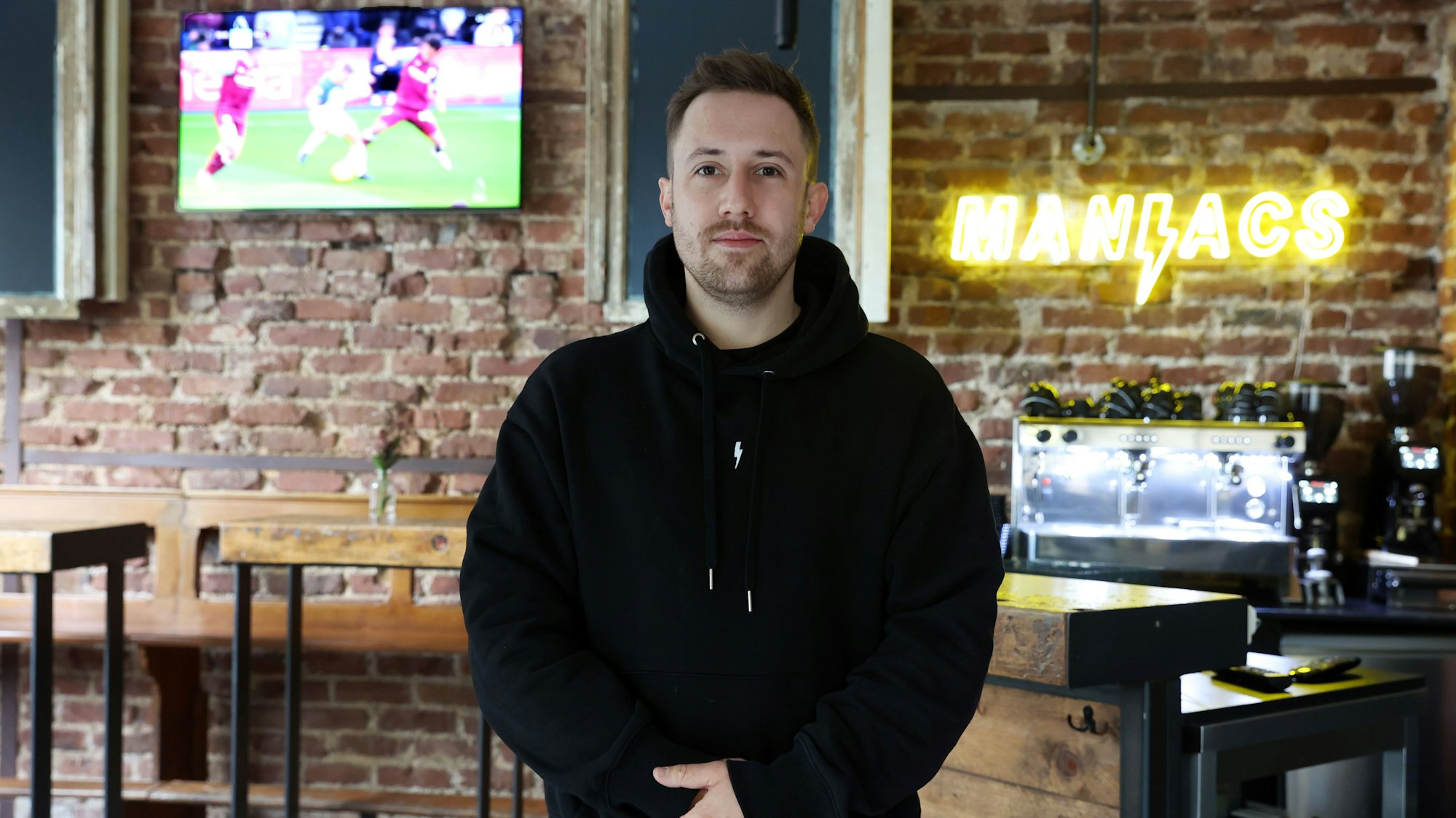 Nicolas Gottschalk, der Wirt der Fußballkneipe Brüsseler dem Brüsseler Platz, steht im Gastraum vor der Theke mit verschränkten Händen. An der Wand hängt ein Bildschirm, auf dem ein Fußballspiel gezeigt wird.