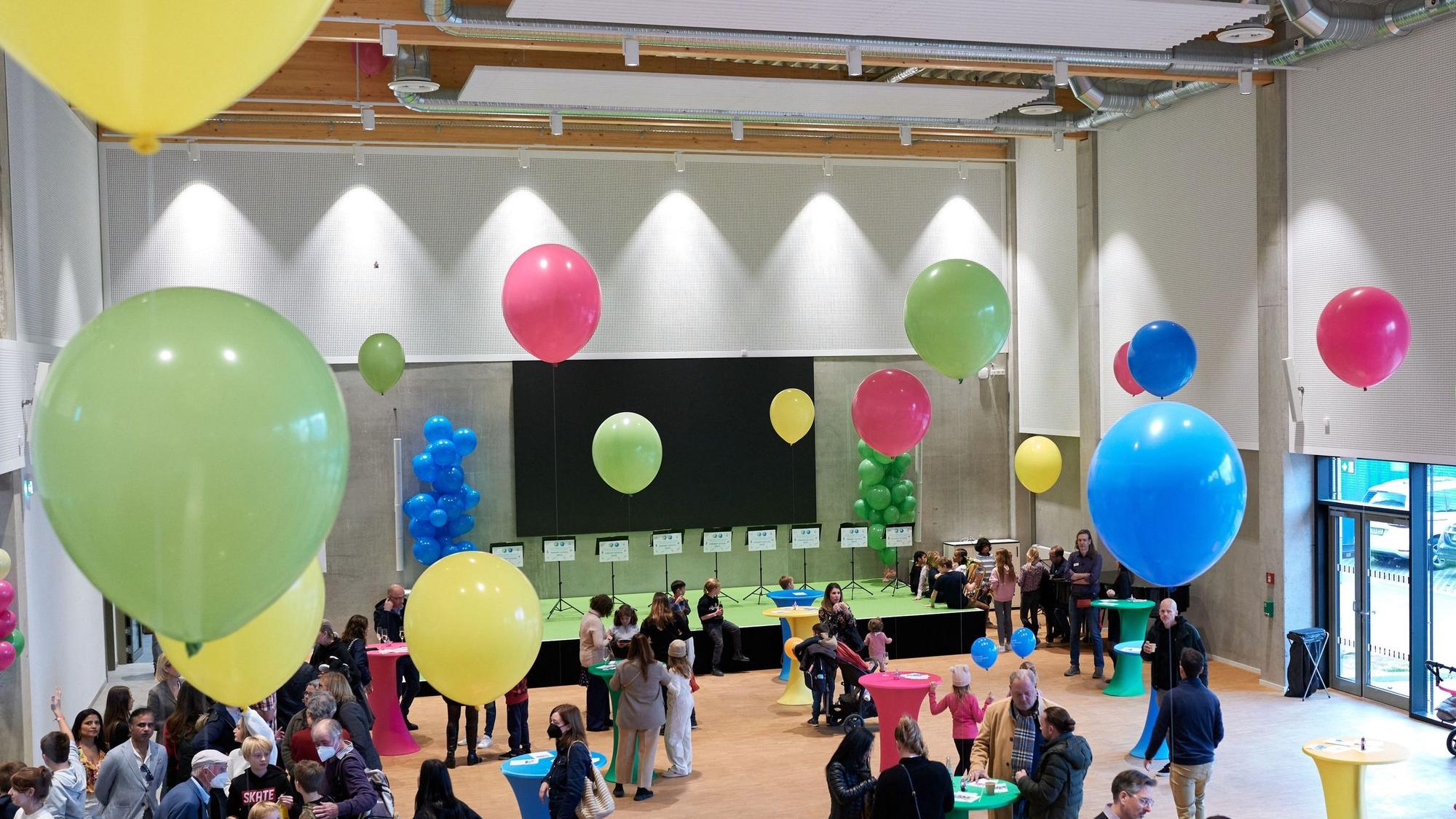 Die zentrale Aula und Multifunktionshalle der Kölner Schule füllt sich mit Eltern und Angehörigen der Schüler. Die Menschen stehen um Stehtische herum. Bunte aufgestiegene Luftballons sind an den Tischen befestigt.