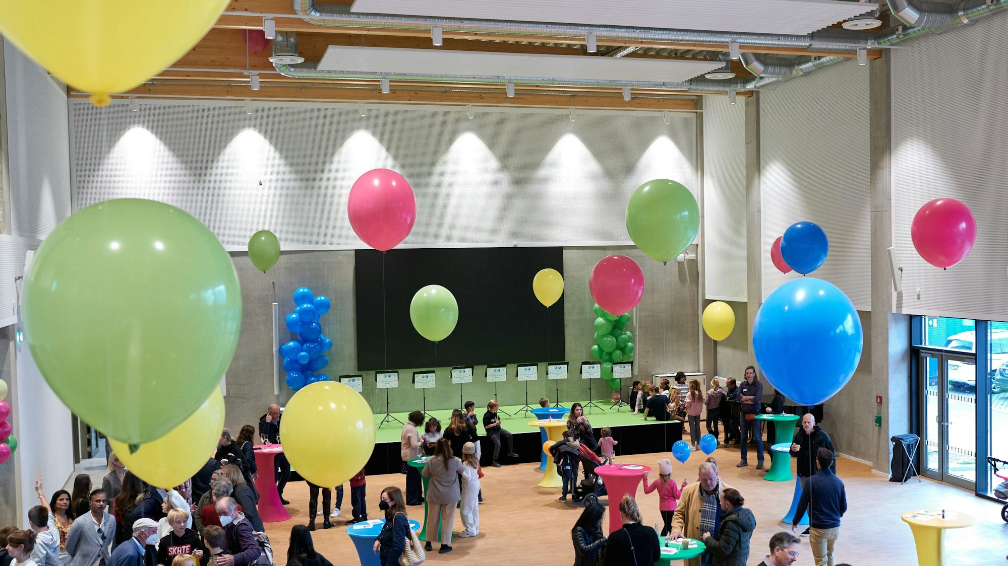 Die zentrale Aula und Multifunktionshalle der Kölner Schule füllt sich mit Eltern und Angehörigen der Schüler. Die Menschen stehen um Stehtische herum. Bunte aufgestiegene Luftballons sind an den Tischen befestigt.