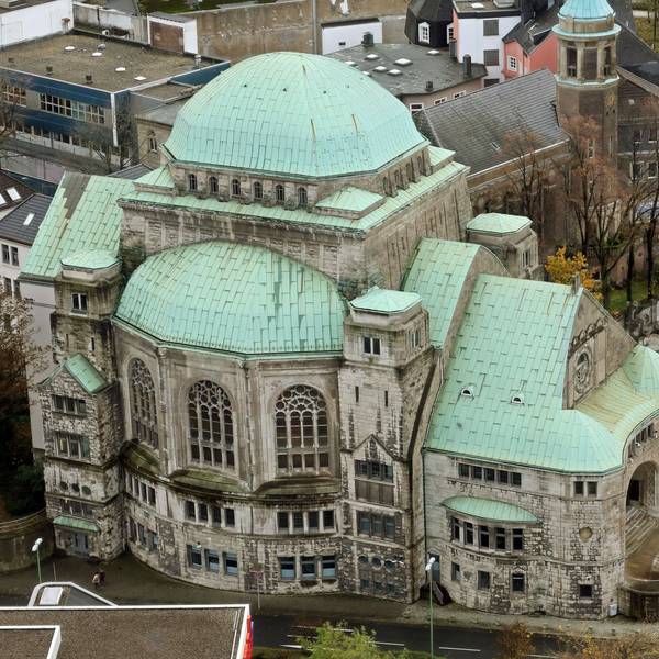 Die Alte Synagoge in Essen ist eine der größten freistehenden Synagogenbauten Europas. Auf dem Bild ist der aufwändige Bau von oben zu sehen, er steht an einer vierspurigen Straße, davor steht ein Polizeiwagen. Das Dach der Alten Synagoge ist mit Grünspan überzogen, die Mauern sind aus hellem Stein.