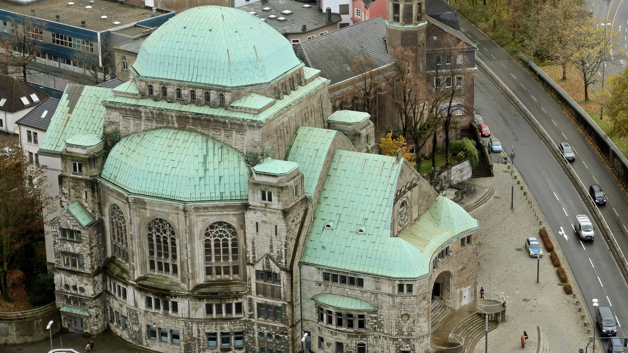 Die Alte Synagoge in Essen ist eine der größten freistehenden Synagogenbauten Europas. Auf dem Bild ist der aufwändige Bau von oben zu sehen, er steht an einer vierspurigen Straße, davor steht ein Polizeiwagen. Das Dach der Alten Synagoge ist mit Grünspan überzogen, die Mauern sind aus hellem Stein.