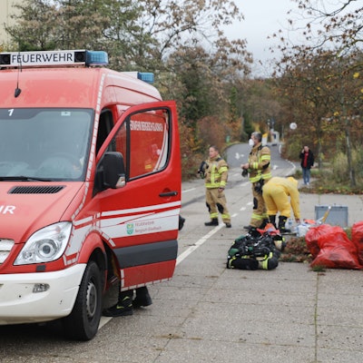Im Vordergrund steht ein kleineres Einsatzfahrzeug der Feuerwehr, im Hintergrund stehen Feuerwehrleute, einer mit einer Schutzmaske und ein weiterer ist mit Schutzbekleidung bekleidet. An der Seite liegen mehrere rote Säcke