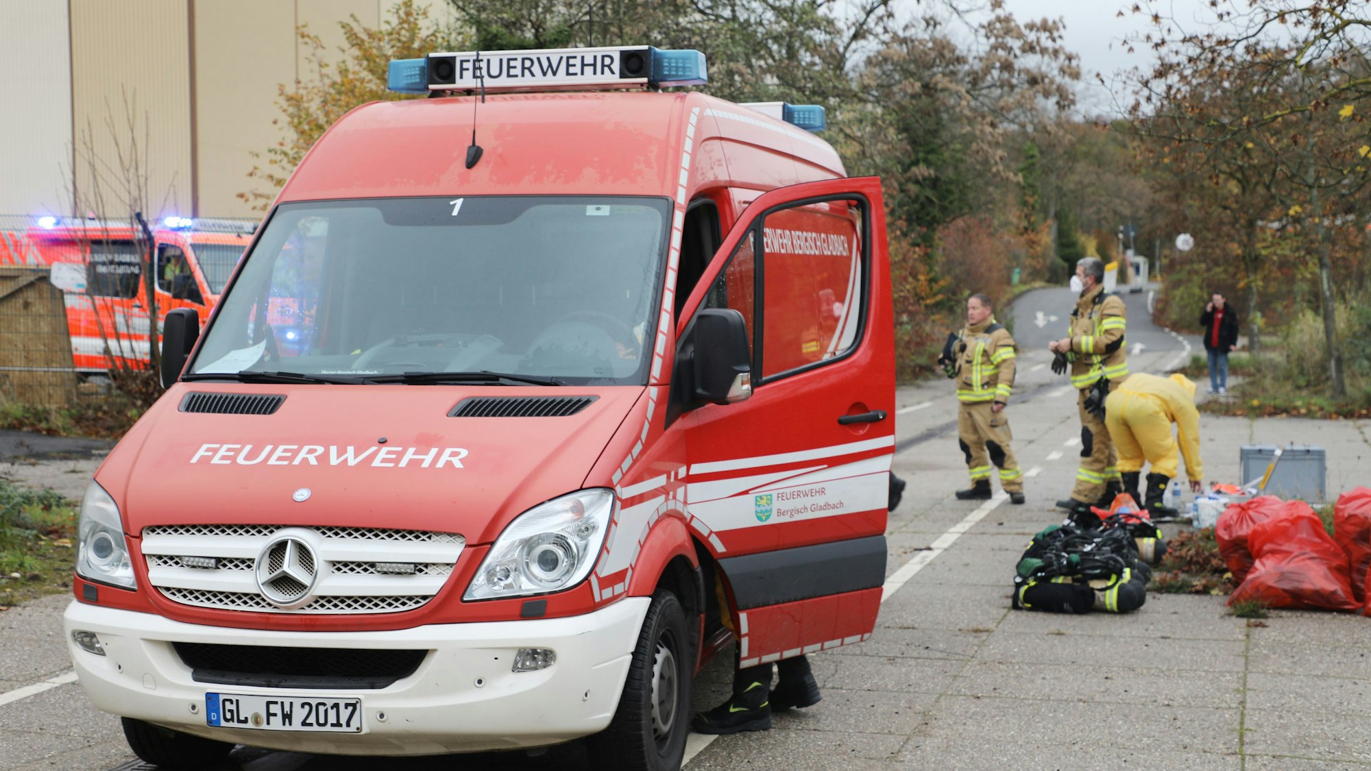 Im Vordergrund steht ein kleineres Einsatzfahrzeug der Feuerwehr, im Hintergrund stehen Feuerwehrleute, einer mit einer Schutzmaske und ein weiterer ist mit Schutzbekleidung bekleidet. An der Seite liegen mehrere rote Säcke